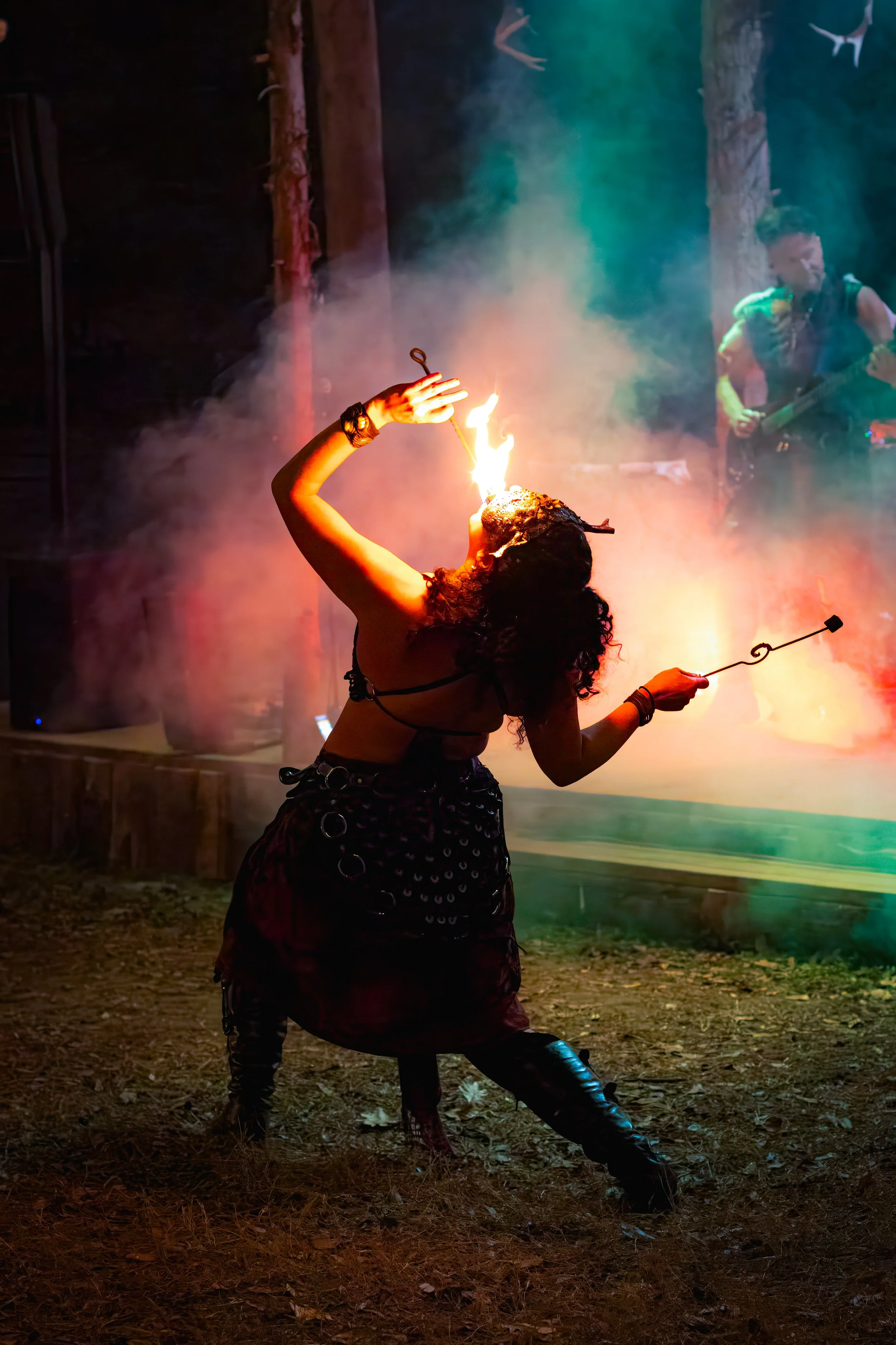 Person performing a fire act with flames on their face during a street performance at night, with musicians playing in the background and colorful smoke.