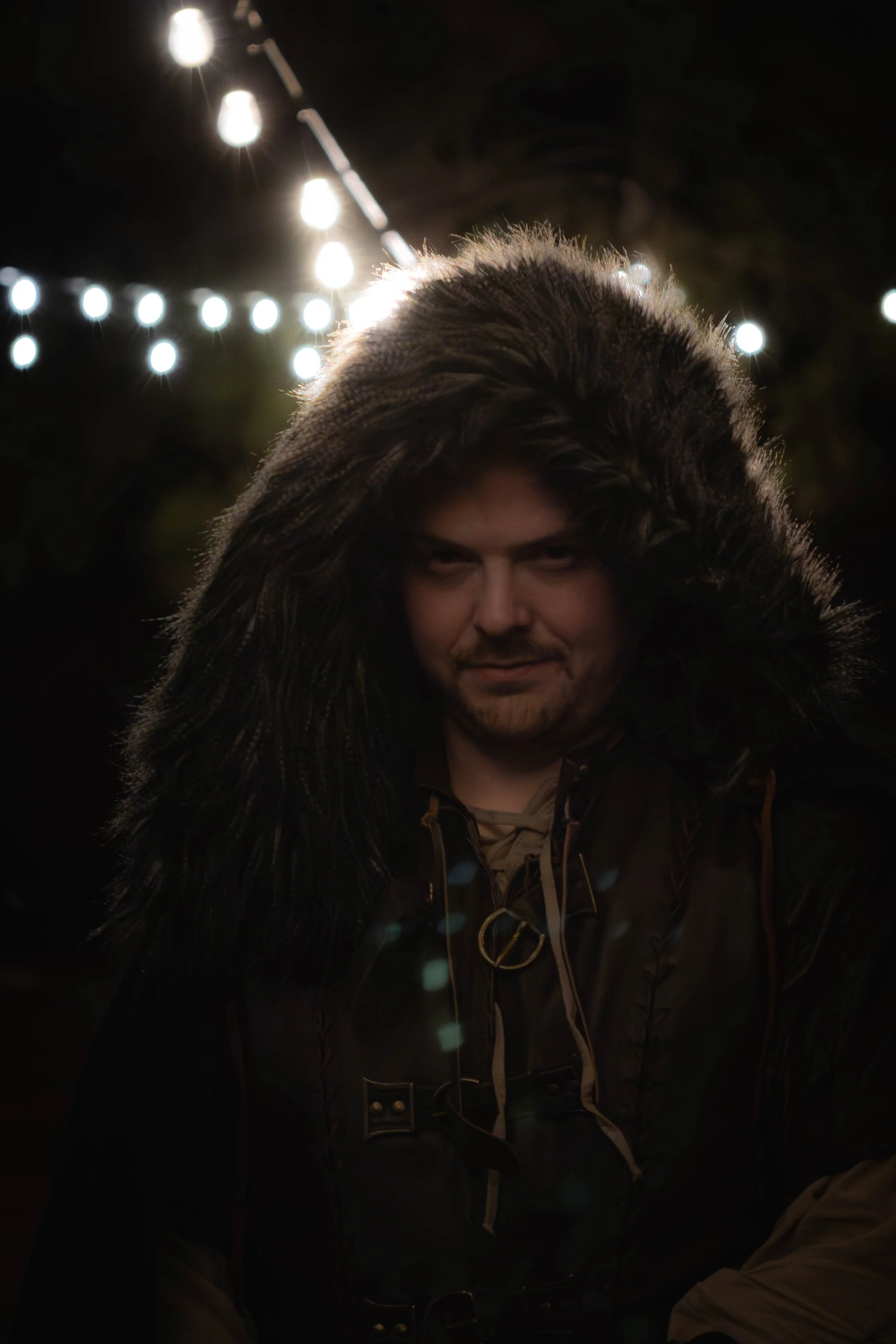 A man wearing a dark coat with a large fur-lined hood, illuminated by string lights in the background at night.