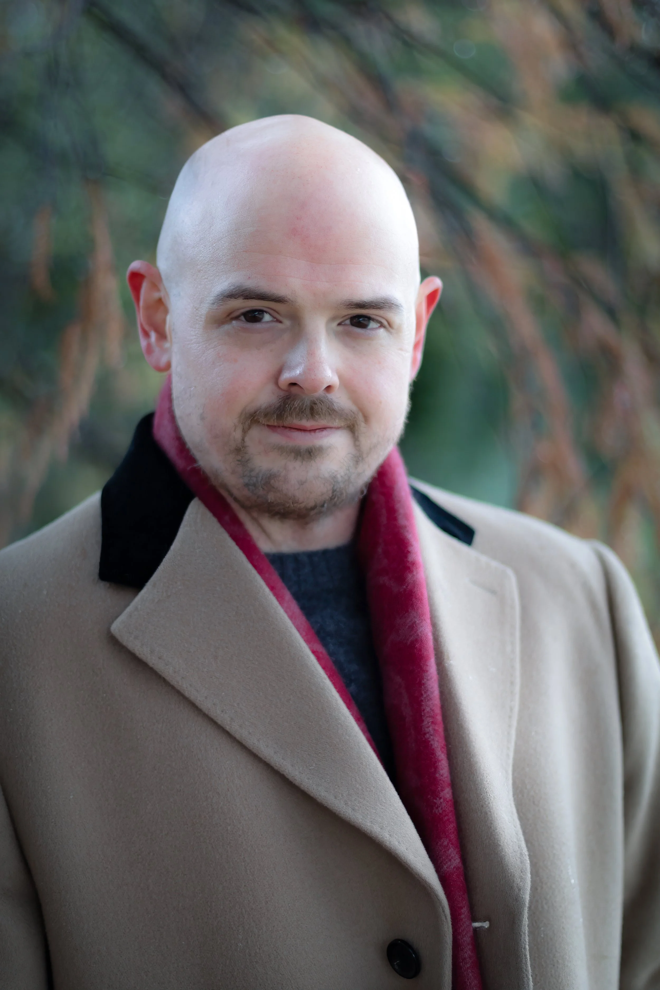 A bald man with a beard and mustache wearing a beige coat with black collar accents and a red scarf, standing outdoors with blurred trees in the background.