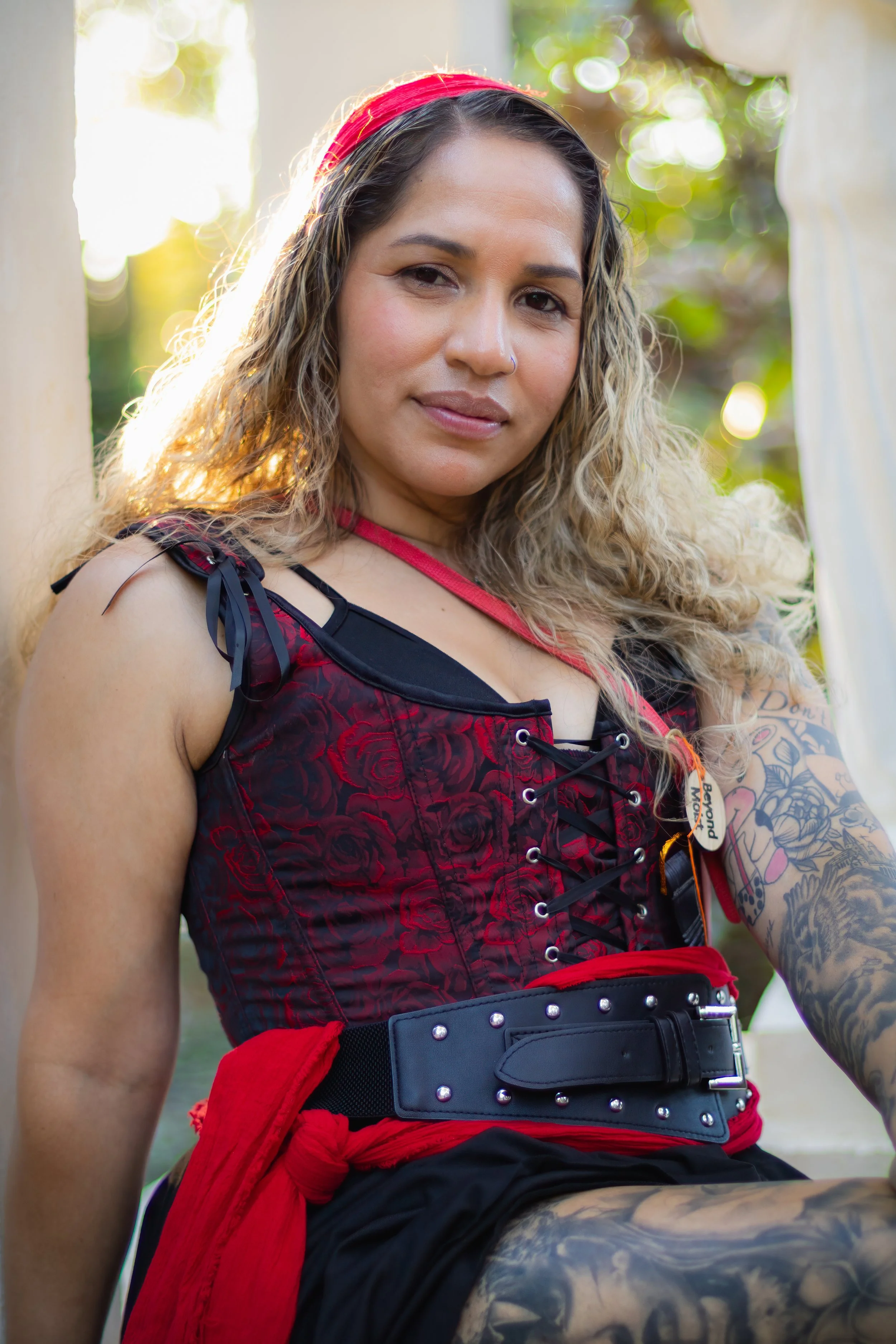 A woman with wavy blonde hair, wearing a red headband, red corset, black skirt, and tattoos on her arm, sitting outdoors near a white structure with green foliage and sunlight in the background.