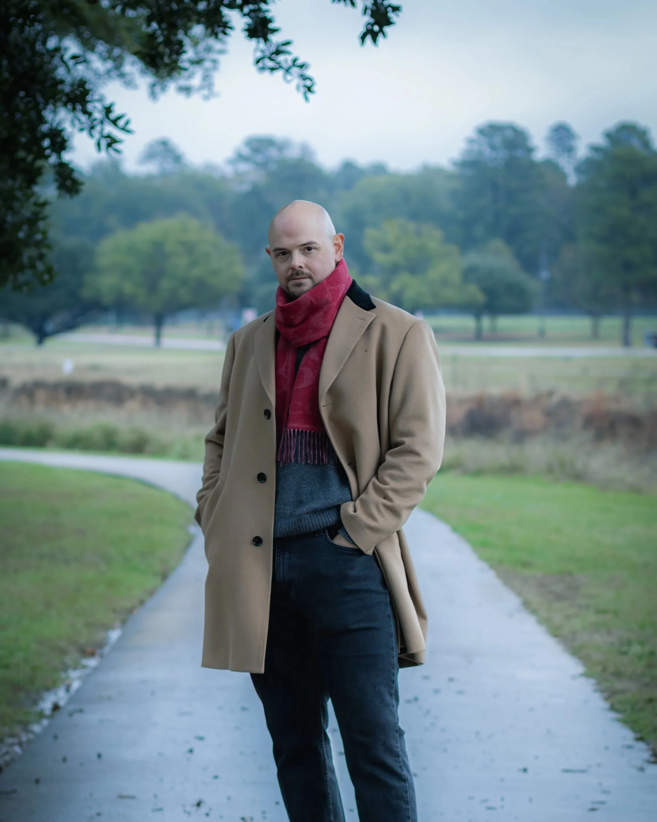 A man standing on a curved pathway in a park, wearing a beige coat, red scarf, and dark jeans, with trees and grass in the background on a cloudy day.