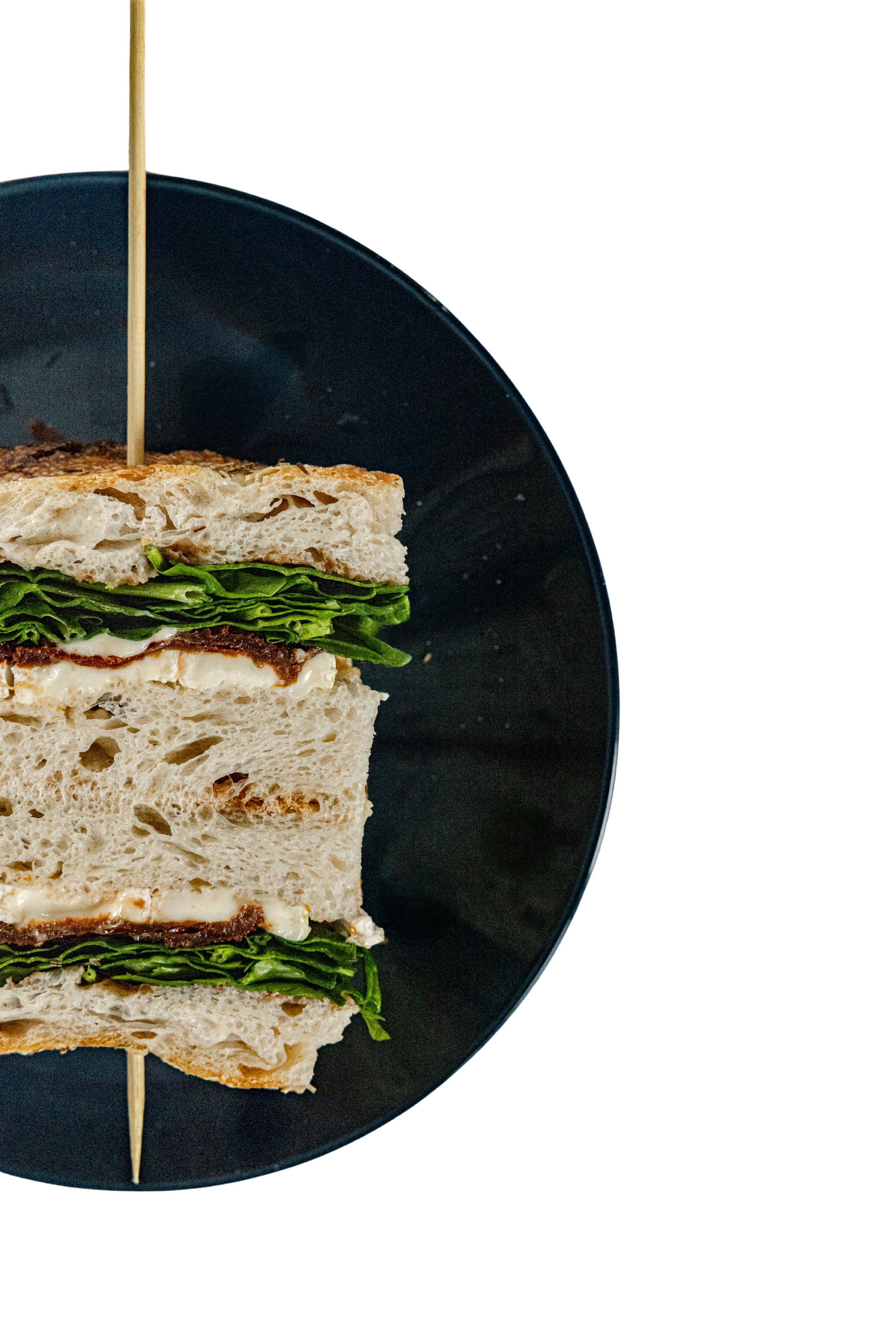 A sliced sandwich layered with leafy greens, cheese, and meat, assembled on bread and skewered with a wooden stick, served on a dark plate.