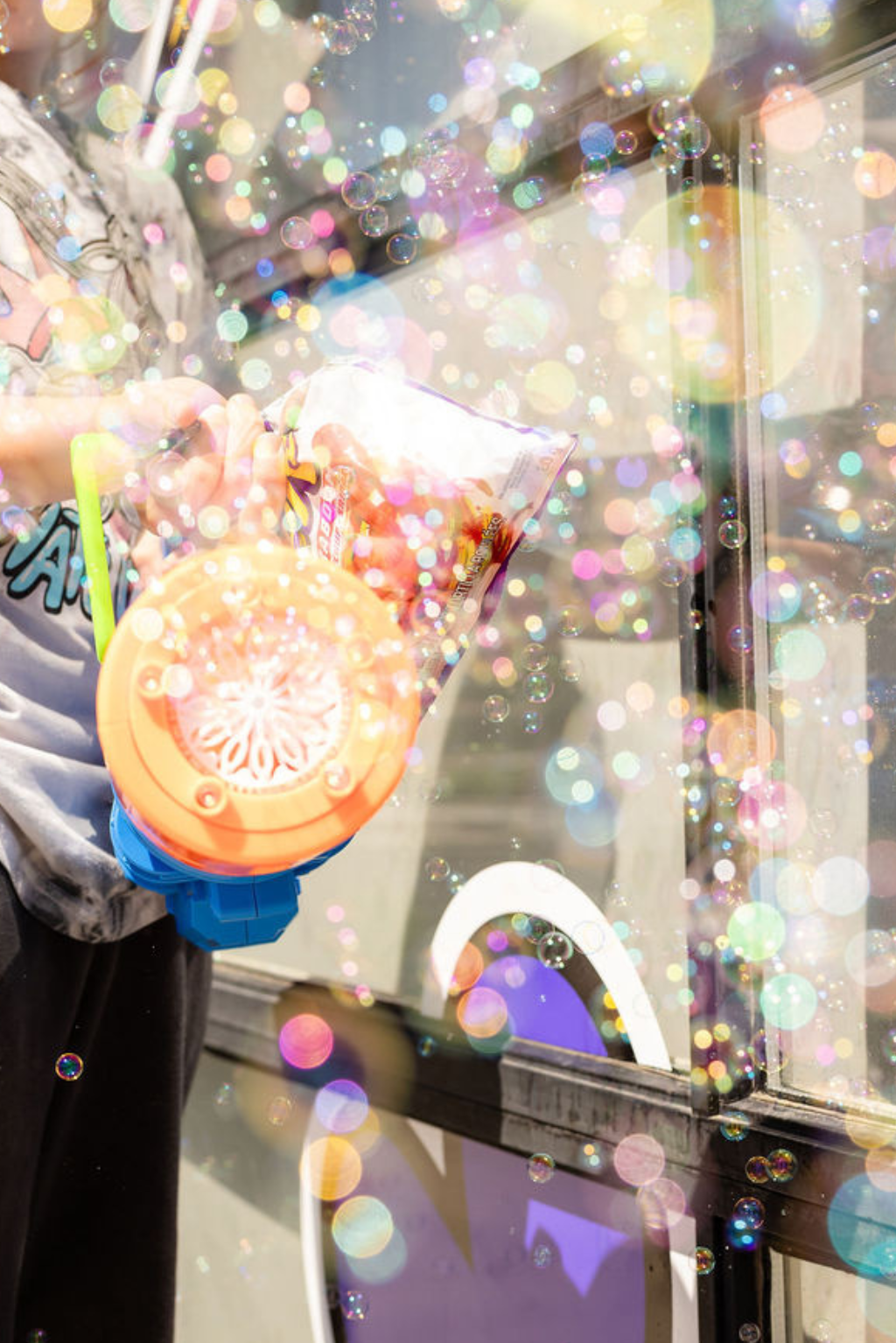 Person holding a pink bubble gun, blowing bubbles outside near a storefront window with colorful reflections and bubbles floating in the air.