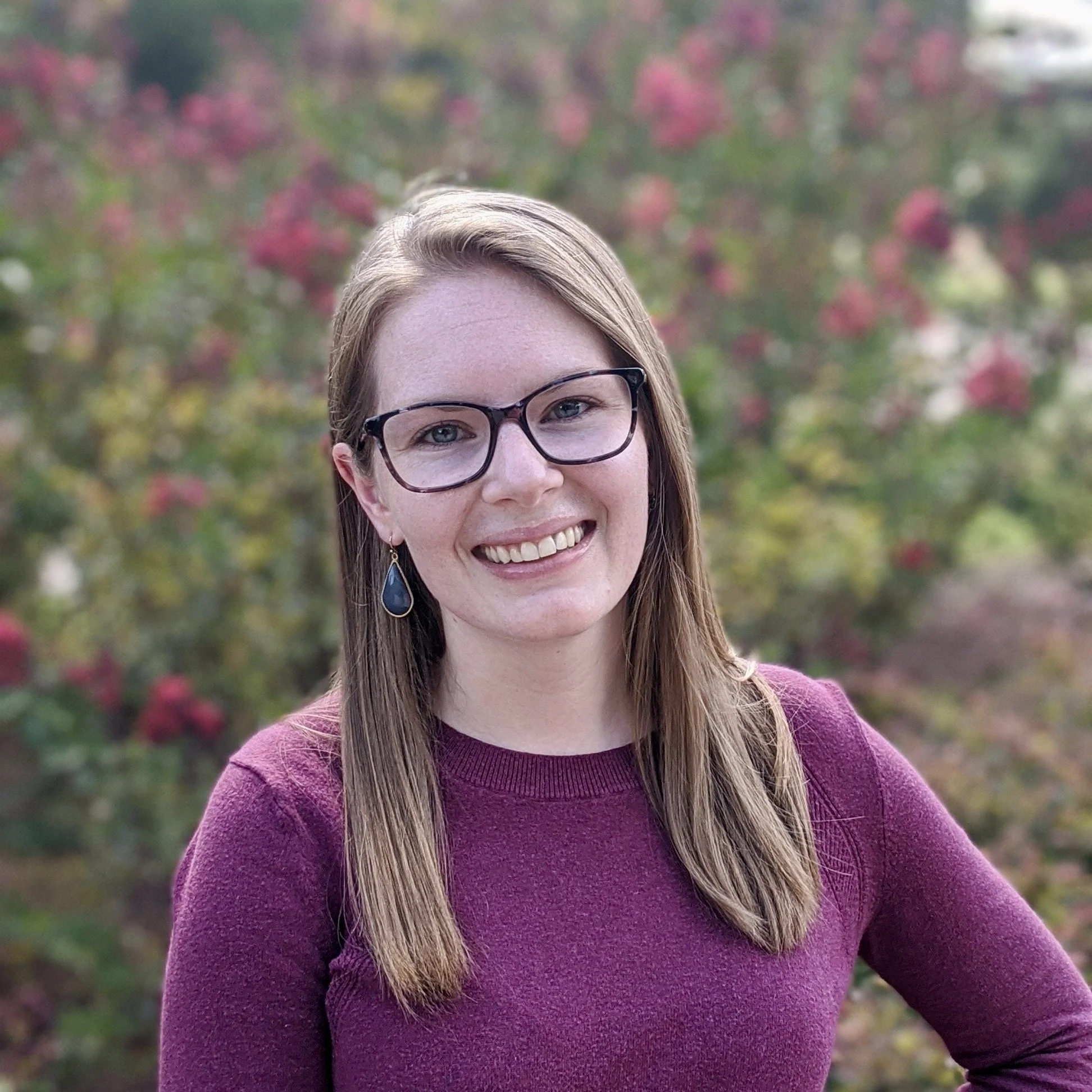 A woman with glasses, long brown hair, wearing earrings and a maroon sweater, smiling outdoors with pink and green foliage in the background.