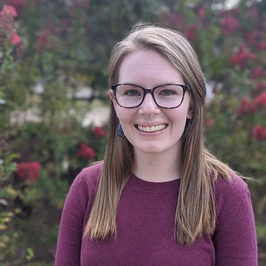 A woman with glasses smiling outdoors, wearing a purple sweater and earrings, with trees and flowers in the background.