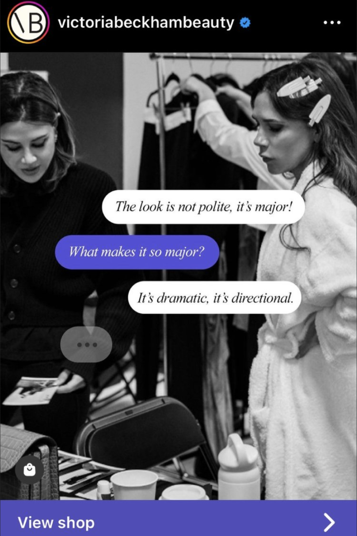 Black and white photo of two women in a behind-the-scenes setting, having a conversation. One woman is holding a photo, and the other has hair clips in her hair. There are clothing racks, a chair, and various objects on a table in the background.