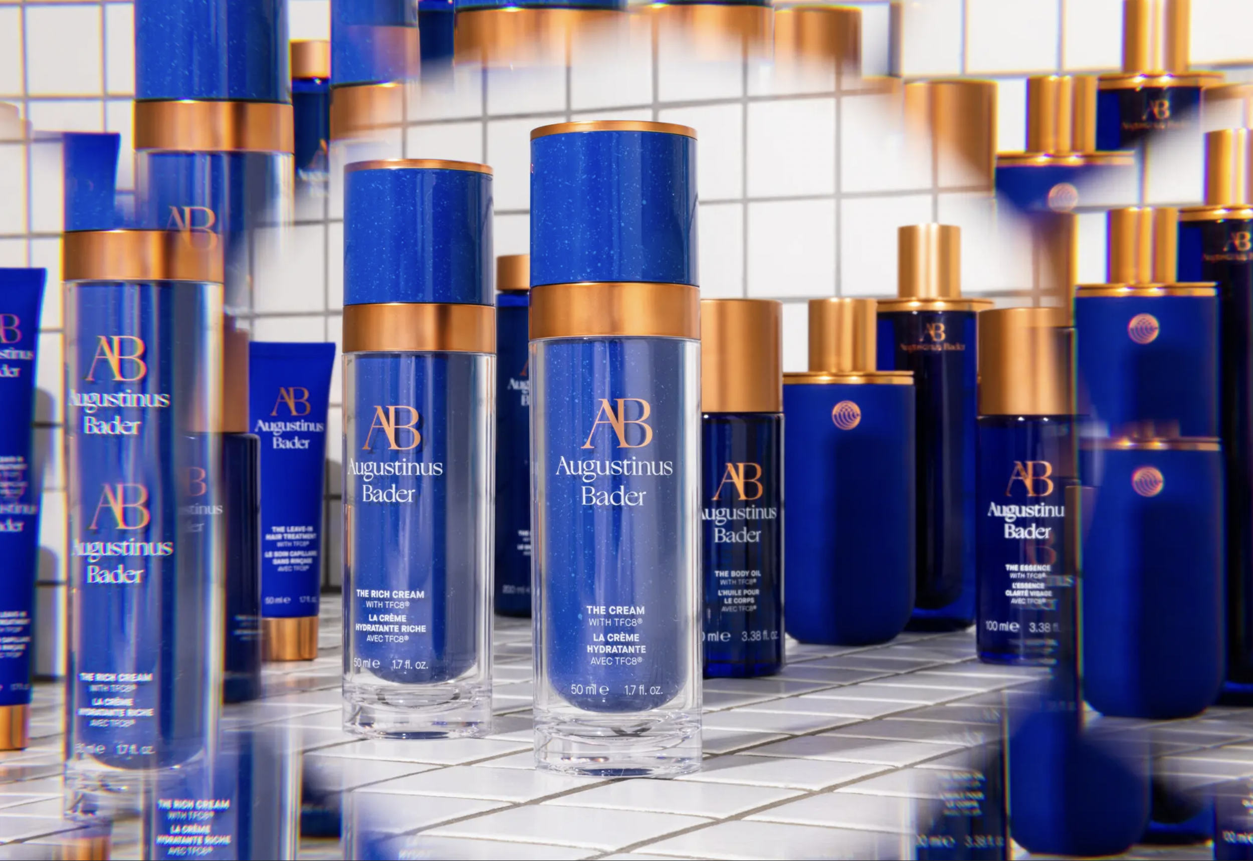 Display of Augustus Bader skincare products on a white tiled shelf, featuring blue bottles with gold caps.
