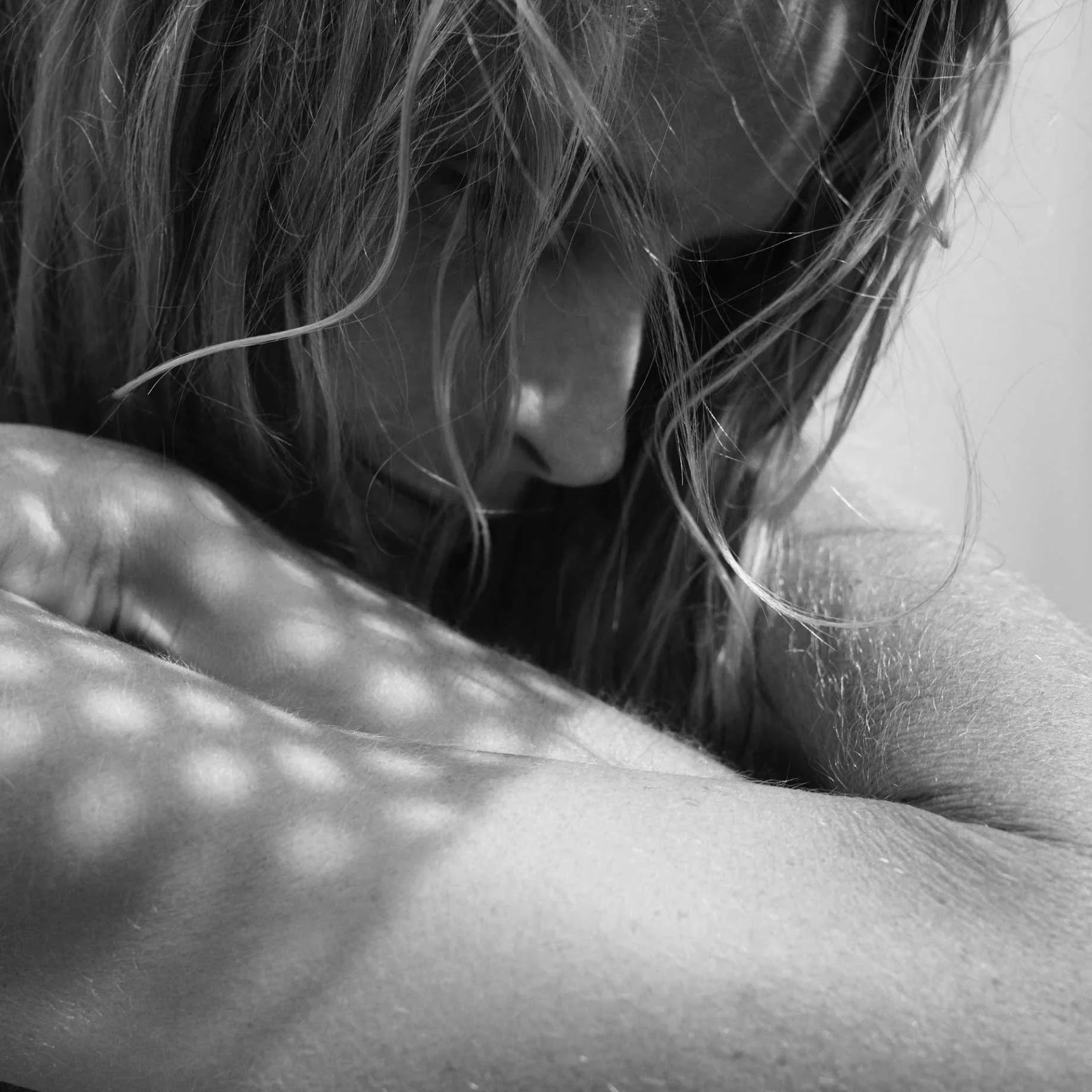 A close-up black and white photo of a woman with wavy hair resting her head on her arm, with a contemplative expression.