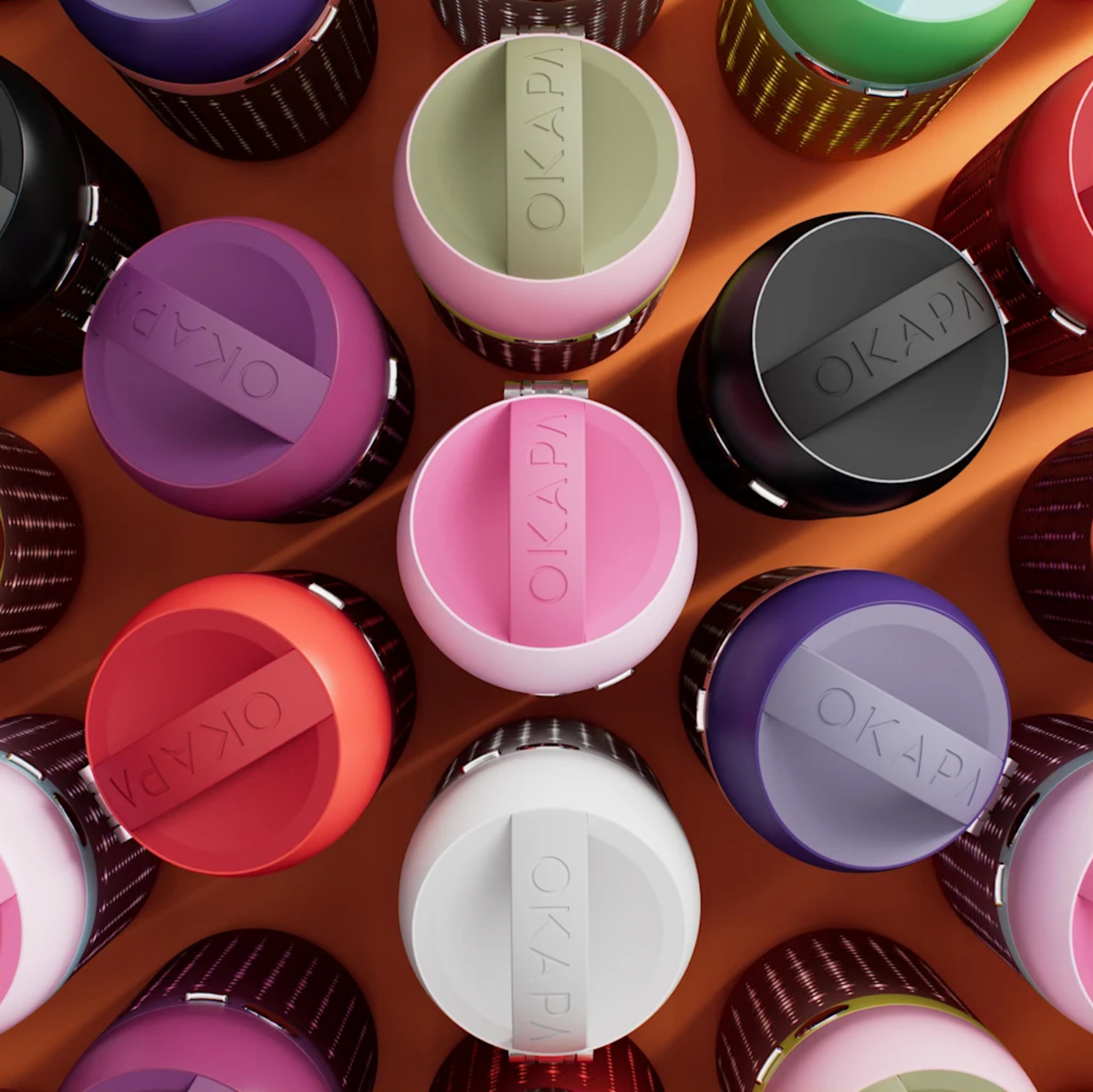 Top-down view of colorful travel mugs with black bases, arranged on a wooden surface. The mugs are in various colors including purple, pink, green, red, white, and gray, with the brand name 'OKAIDI' embossed on the lids.
