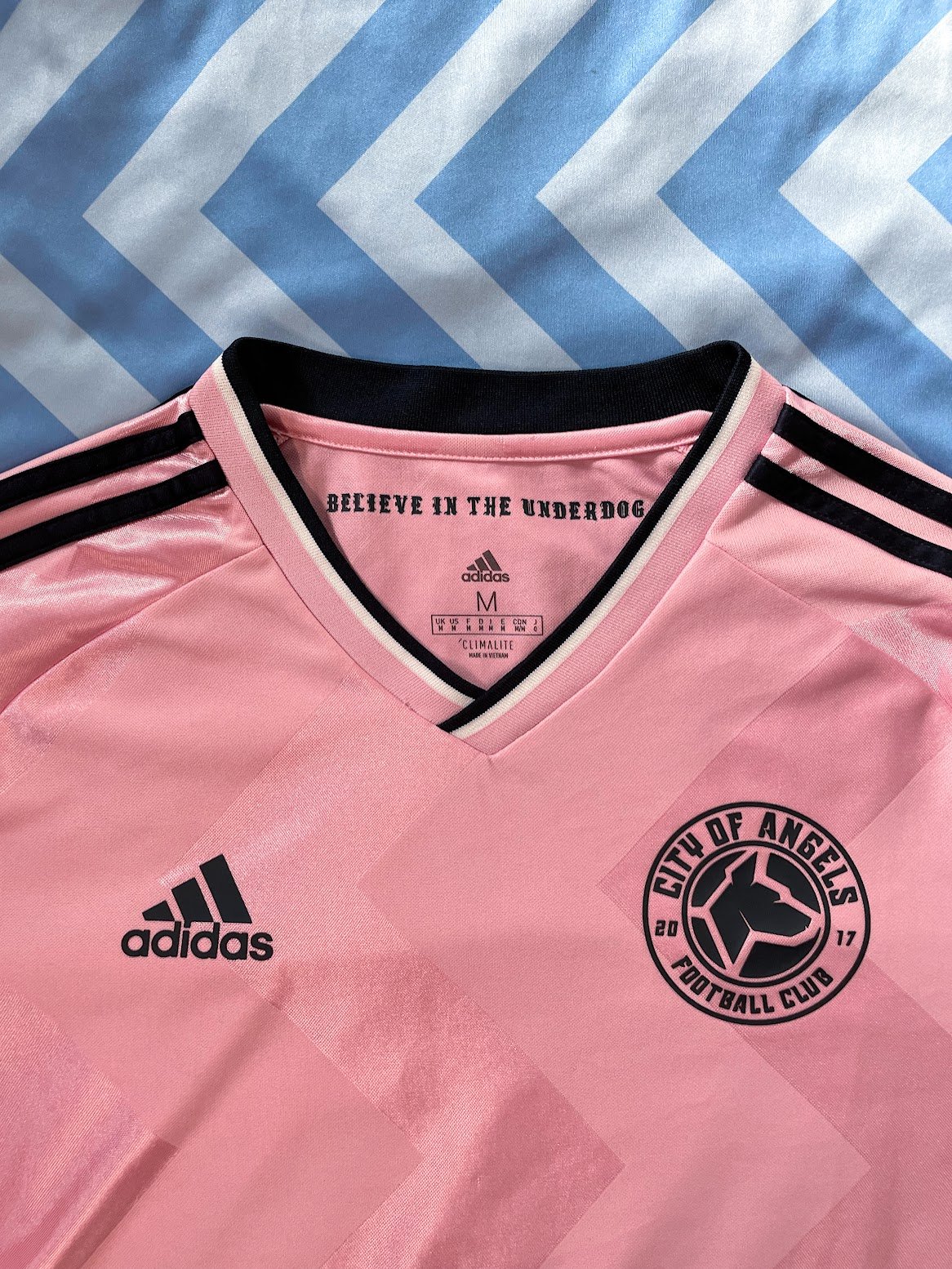 Pink City of Angels Football Club soccer jersey with Adidas logo, black team logo, and black and pink stripe accents, laid on a blue and white chevron-patterned fabric background.