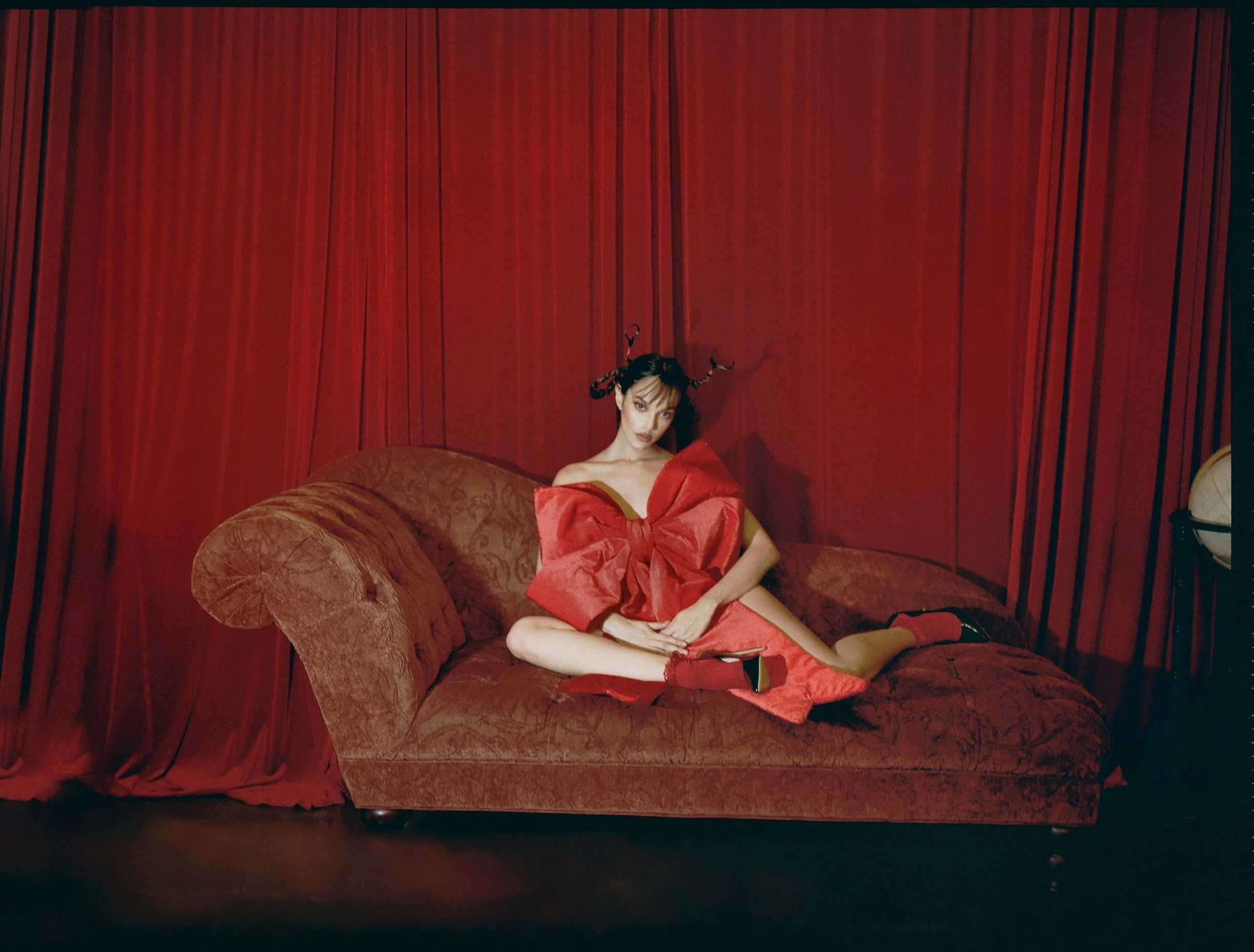 A woman sitting on a red vintage sofa against red curtains, wearing a red dress with exaggerated bow detail, red shoes, and with black hair styled with horns.