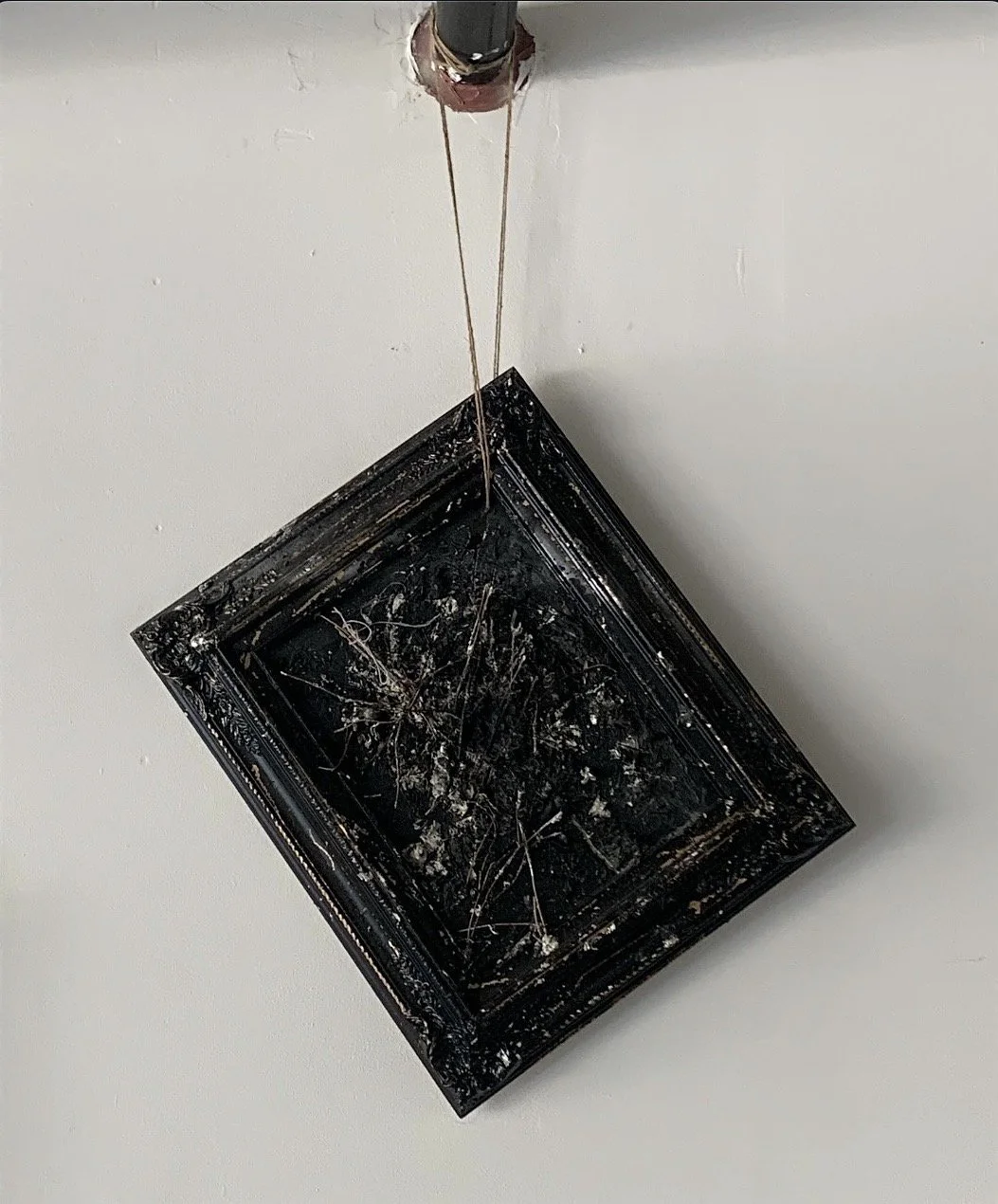 A damaged, black, ornate picture frame hanging upside down on the ceiling with a broken glass panel and spider webs inside.