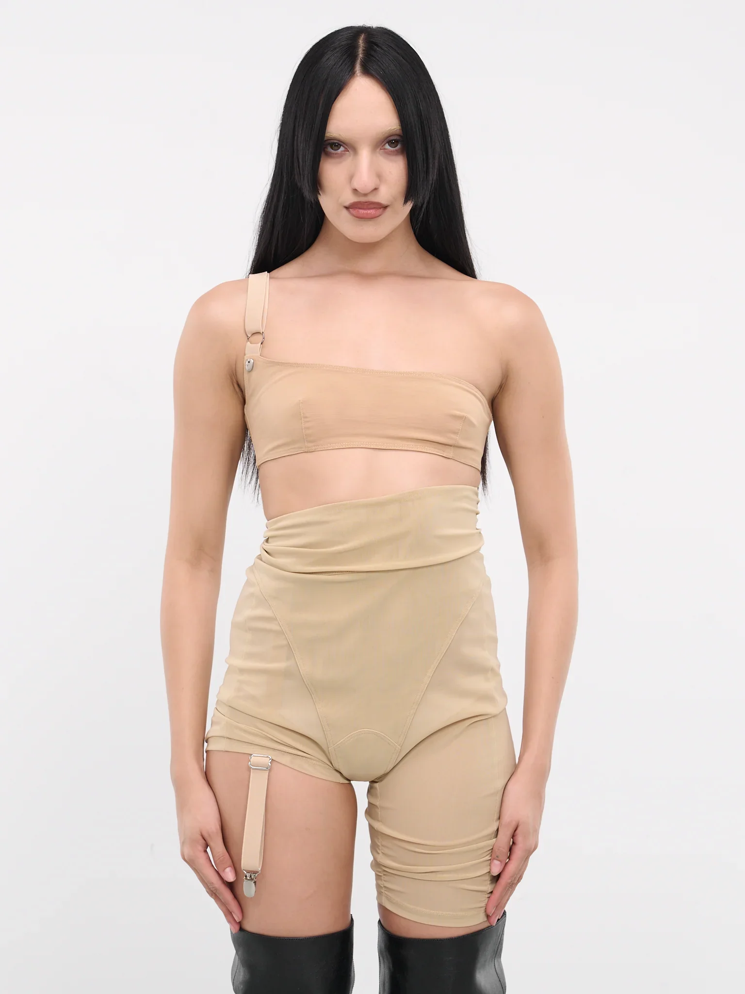 A woman with long black hair wearing a beige asymmetrical top and high-waisted beige shorts with suspenders, standing against a plain white background.