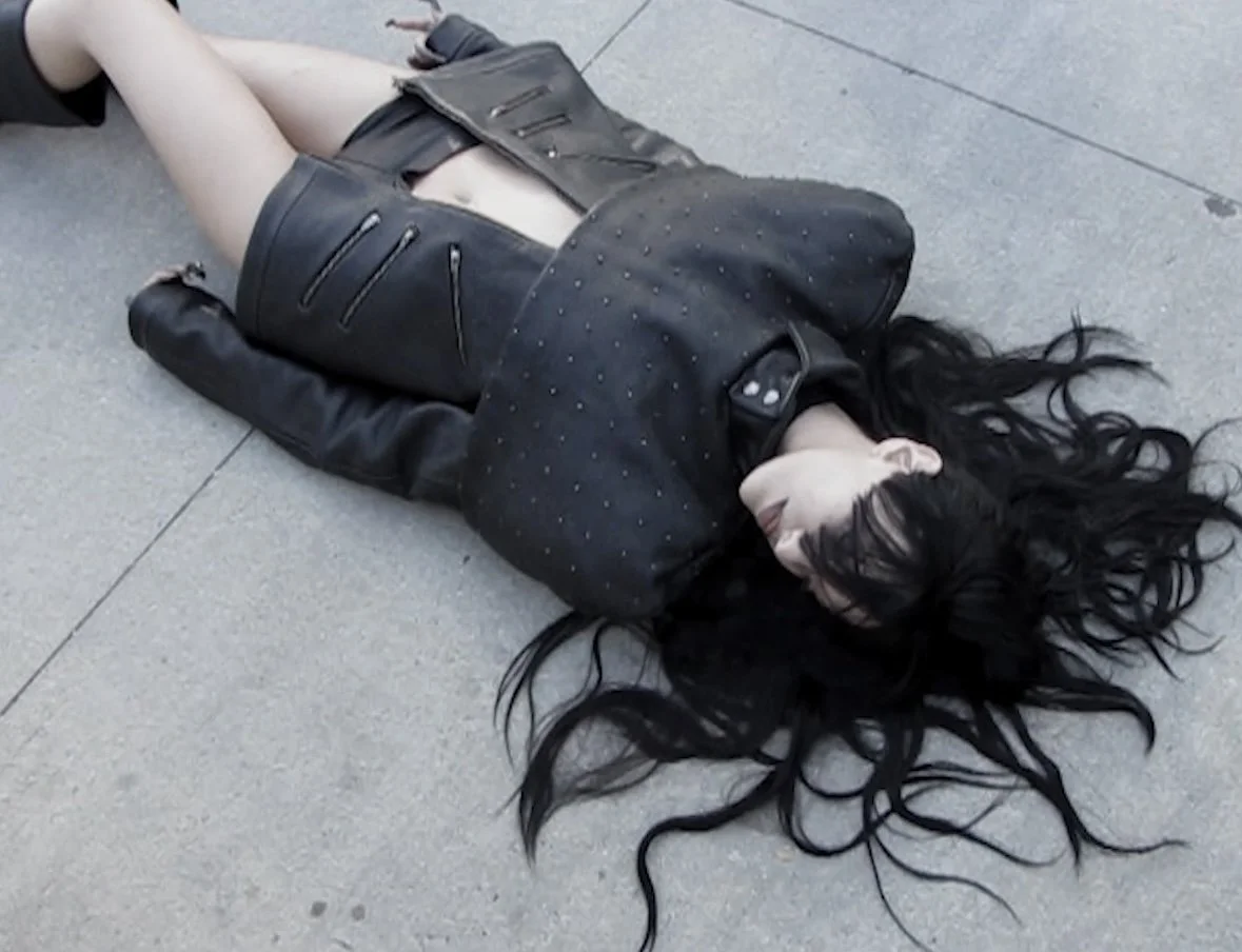 A woman lying on the sidewalk, wearing a black leather jacket and skirt, with long black hair spread out on the ground.