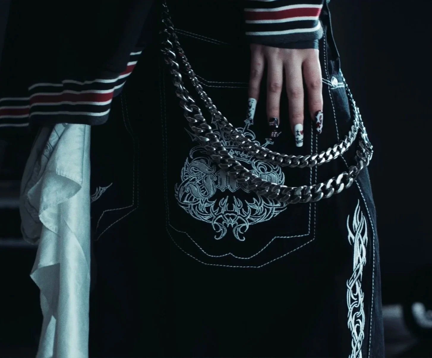 Close-up of a person wearing black jeans with embroidered white designs, a striped long-sleeve shirt, and a chain belt hanging from the waistband. The person's hand with painted nails is resting on the hip.