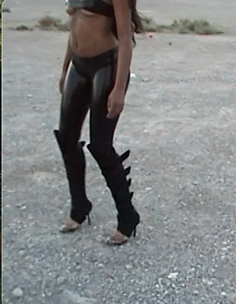 Person wearing shiny black leggings and high-heeled shoes, standing on a gravel surface.