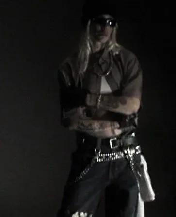 Person with long blonde hair wearing sunglasses, a brown shirt, black pants with chains, and holding a gun, standing in a dark environment.