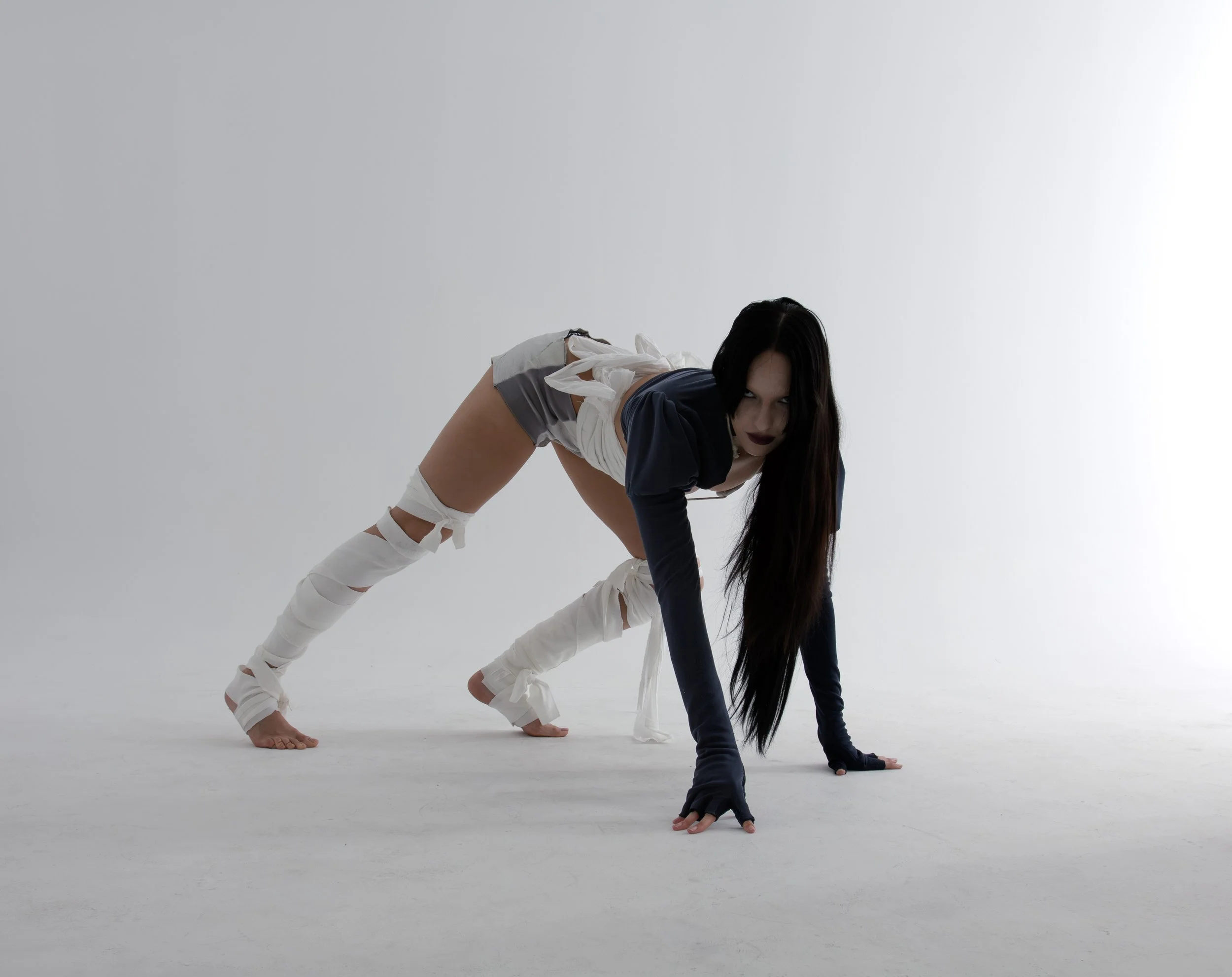 A woman with long dark hair and dark lipstick is in a crawling pose on a white studio floor, wearing a black long-sleeve top, torn gray shorts, and white leg wraps with ties.