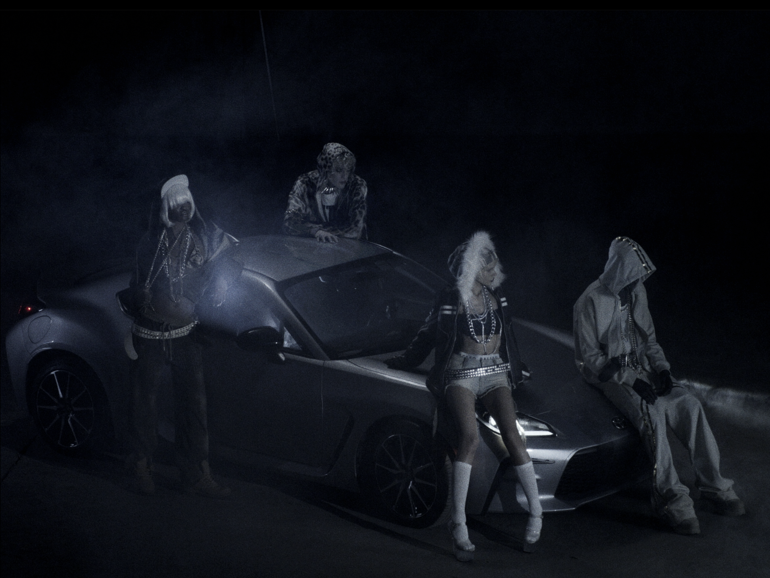Four individuals dressed in fashionable, edgy clothing, some with accessories like chains and hats, gathered around a sleek sports car in a dark, moody setting.
