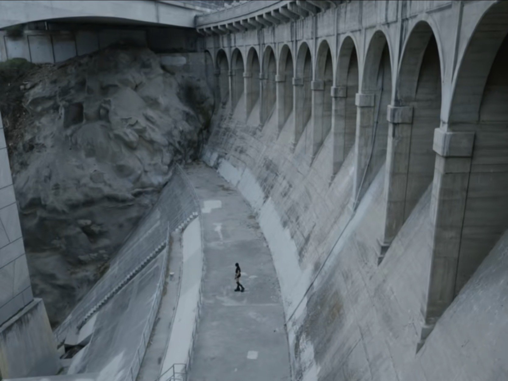 A person standing near the base of a large concrete dam with arched openings, and a rocky hillside in the background.
