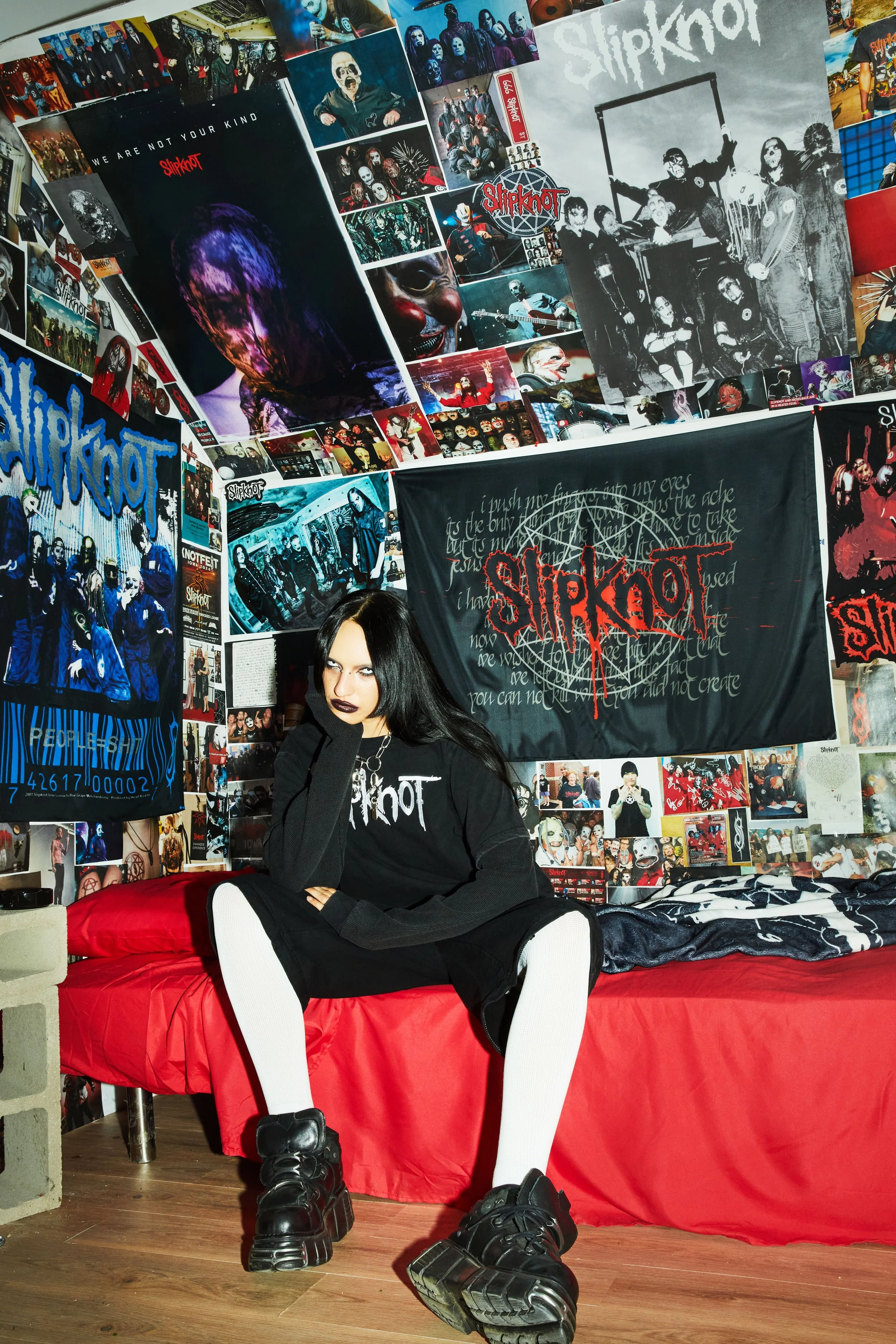 A young woman with dark hair and gothic style makeup sitting on a red bed in a room decorated with Slipknot posters, flags, and photos, all related to the band Slipknot.