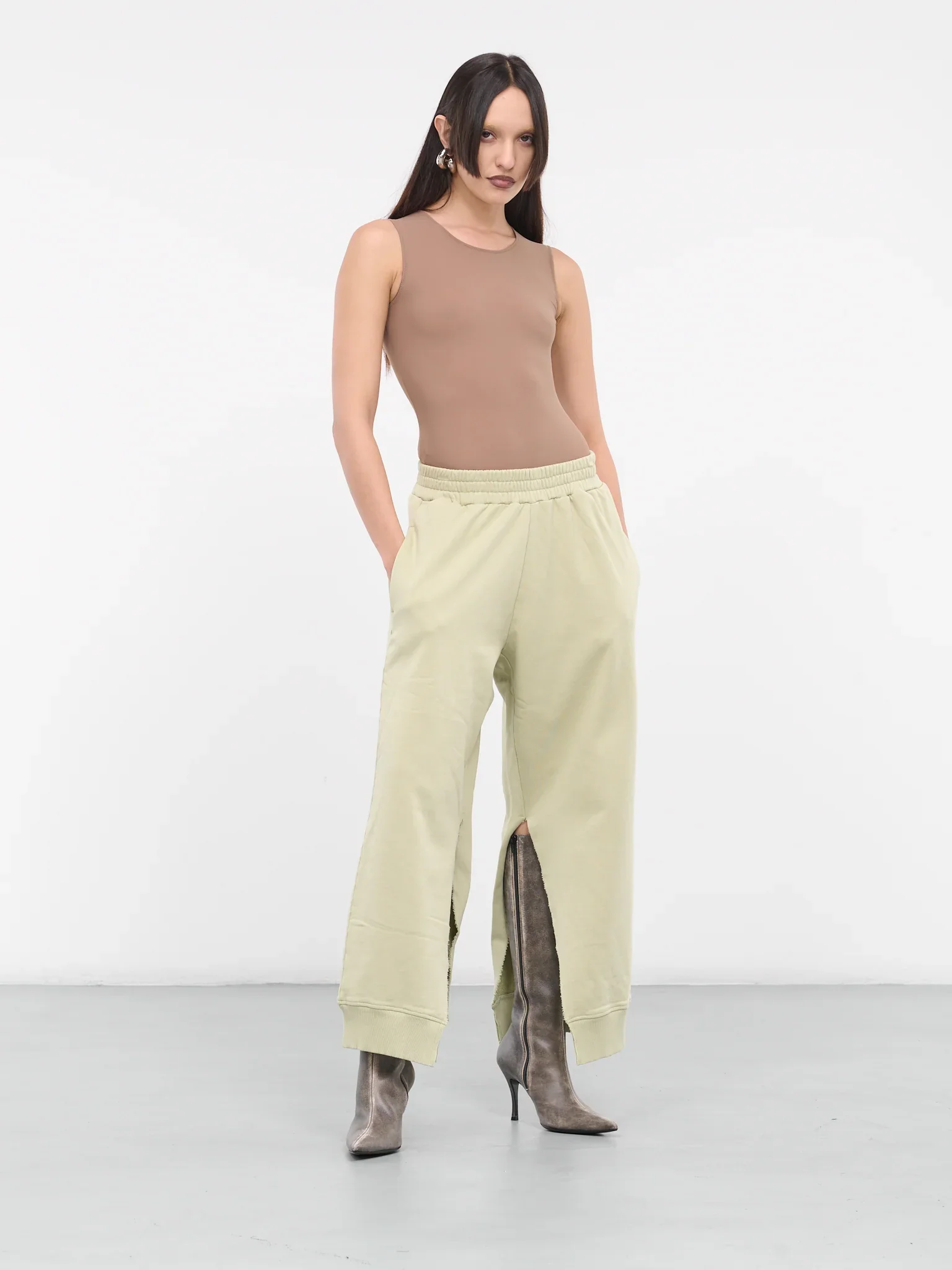 A young woman with long dark hair wearing a beige sleeveless top, light yellow wide-leg pants with slits at the bottom, and gray high-heeled boots, standing with her hands in her pockets against a plain white background.