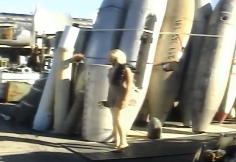 A woman walking past large, weathered missile shells stacked outdoors at a shipyard or military storage area.