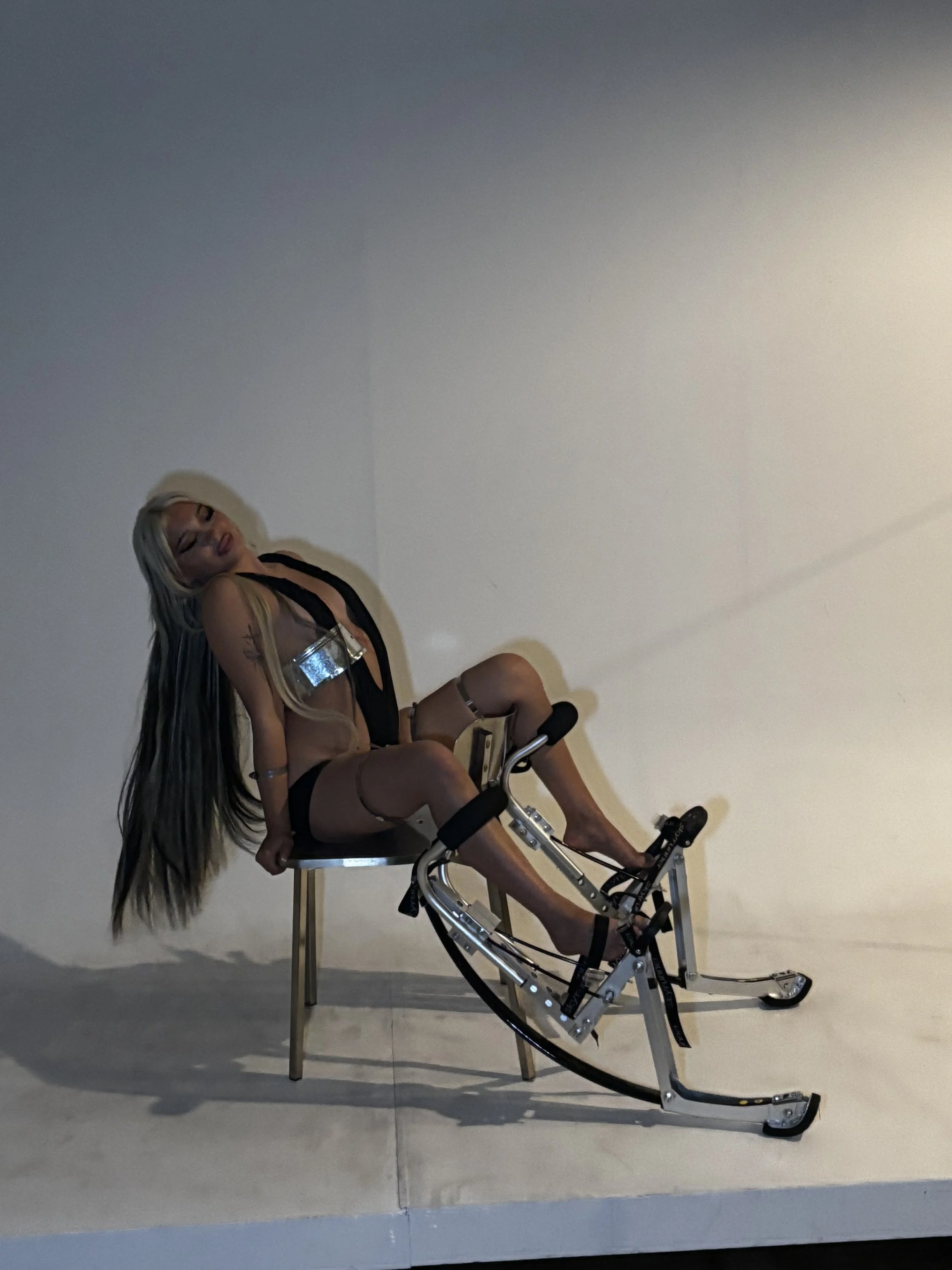 A woman with long blonde hair sits on a black chair, leaning back with her eyes closed. She is wearing a revealing black outfit and is seated on a mechanical device, possibly a rehabilitation or exercise machine, with a beige background.