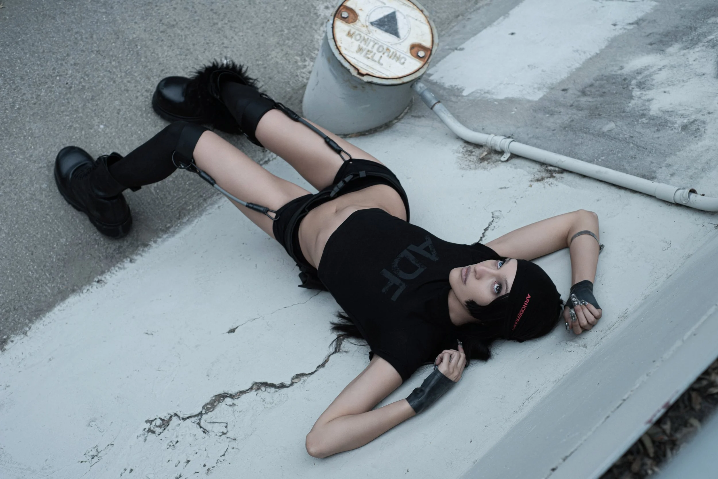 A young woman with black hair, wearing black boots, knee-high socks, black shorts, a black t-shirt, and a black headband, lies on her back on a cracked concrete surface next to a white wall and a metal pipe, looking upward with her right arm bent and
