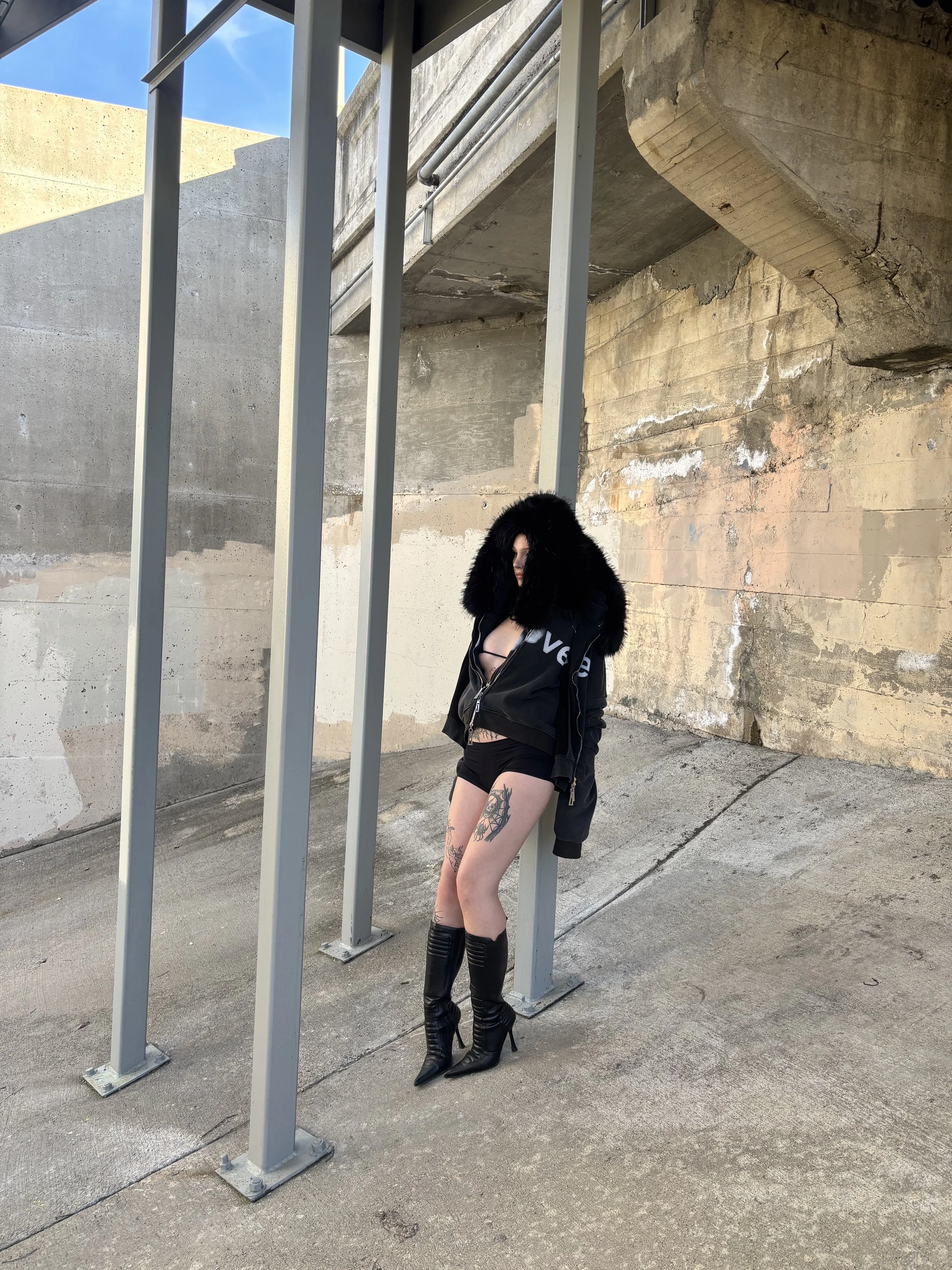 A woman standing under a concrete overpass, wearing black high-heeled boots, black shorts, a black jacket with a fur hood, and a black top. She has tattoos on her thighs and is standing near metal support beams.