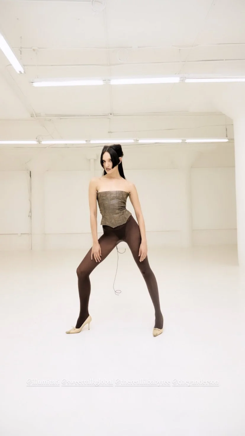 A woman with shoulder-length black hair and pale skin poses in a large, empty, brightly lit white room. She wears a strapless, fitted top, black tights, and beige high heels, standing with her legs apart and hands resting on her thighs.