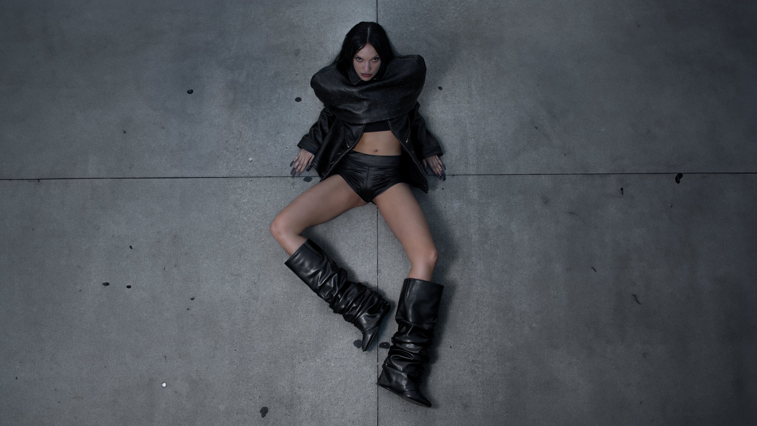 A woman with dark hair lying on a concrete floor, wearing black leather shorts, a leather jacket, and thigh-high black leather boots, looking up at the camera.