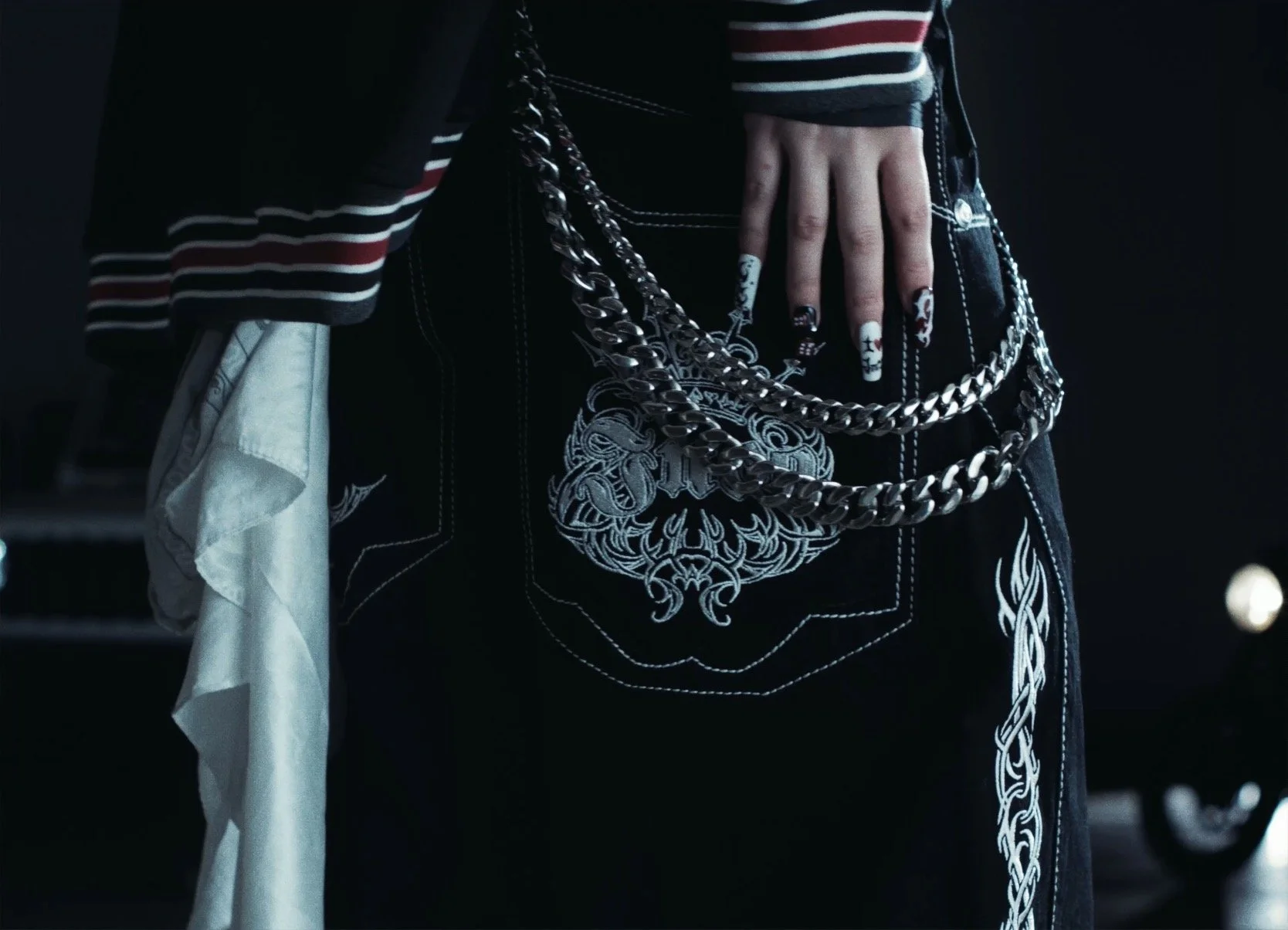 Close-up of a person's hip with a black embroidered design on their dark jeans, multiple silver chains hanging from the belt loop, and a striped sleeve and a white cloth visible on the person.