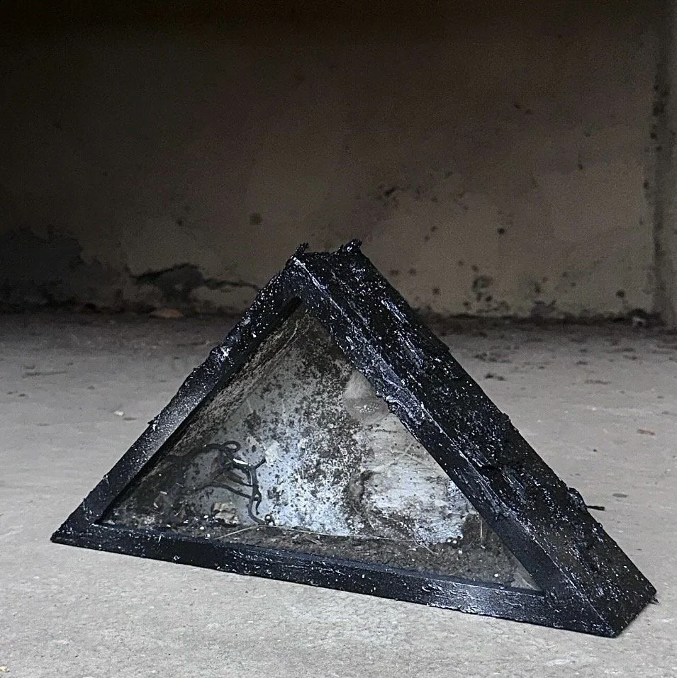 A black metal pyramid-shaped object with a glass front, containing ashes and small metal pieces, placed on a concrete surface with a brick wall background.