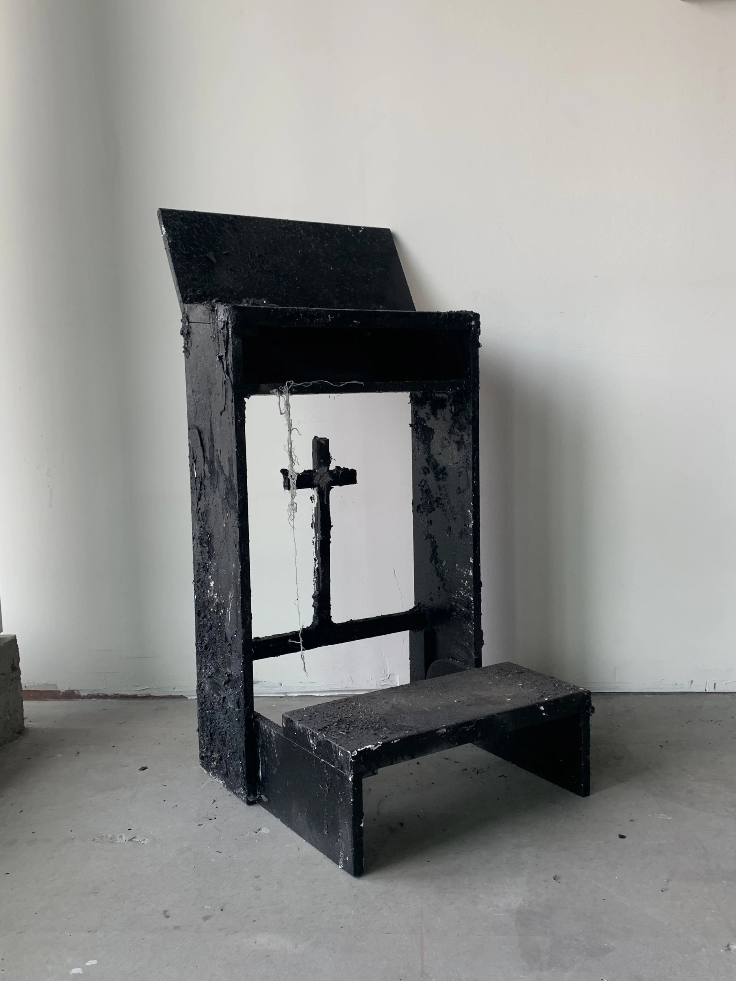 A black cross statue attached to a black altar with a small shelf, made of a rough, possibly burnt or charred material, placed on a concrete floor against a white wall.