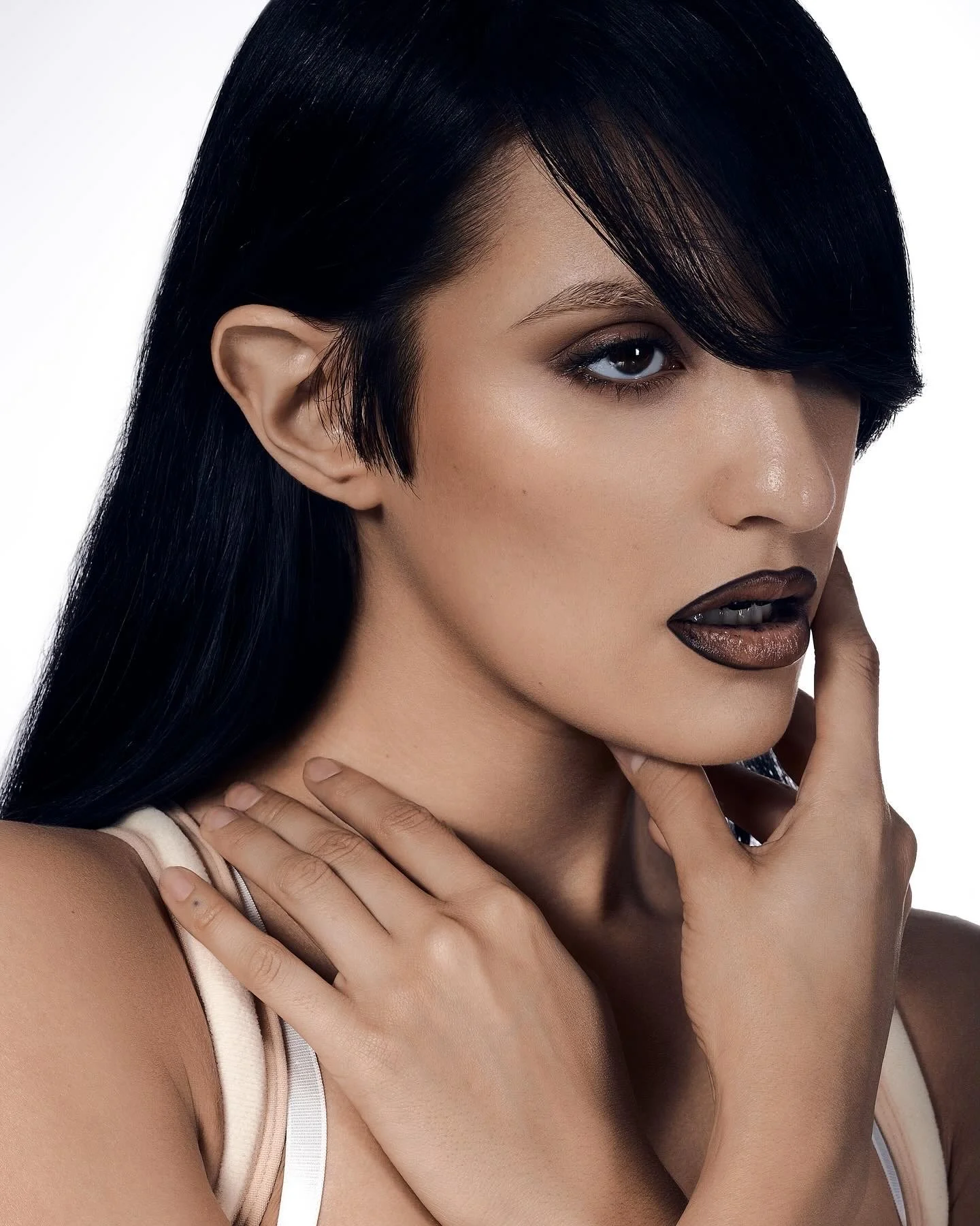 Close-up of a woman with dark hair, smoky eye makeup, and glossy dark lipstick, touching her face with one hand.