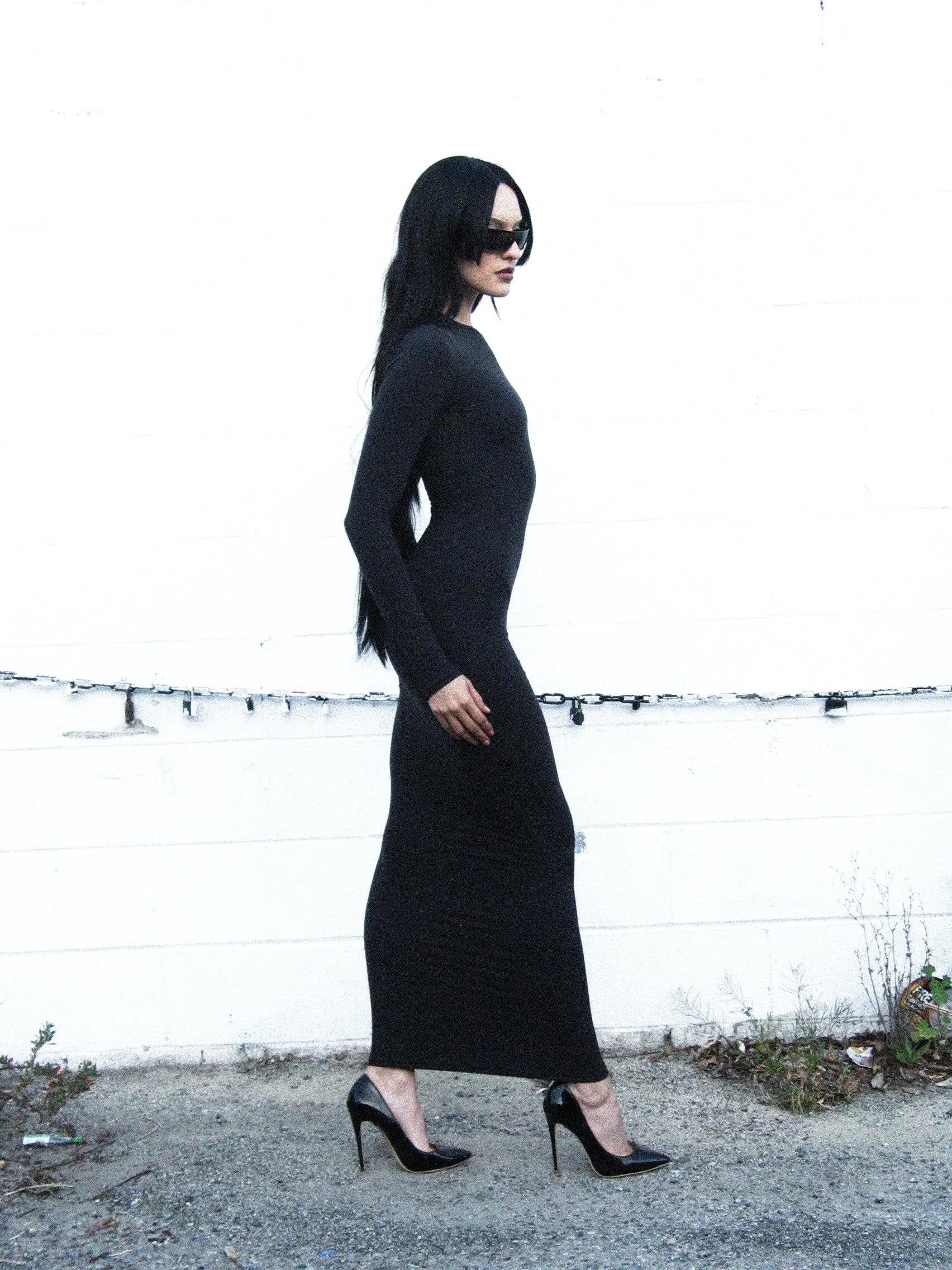 A woman in a long black dress and high heels walking outdoors against a white wall.