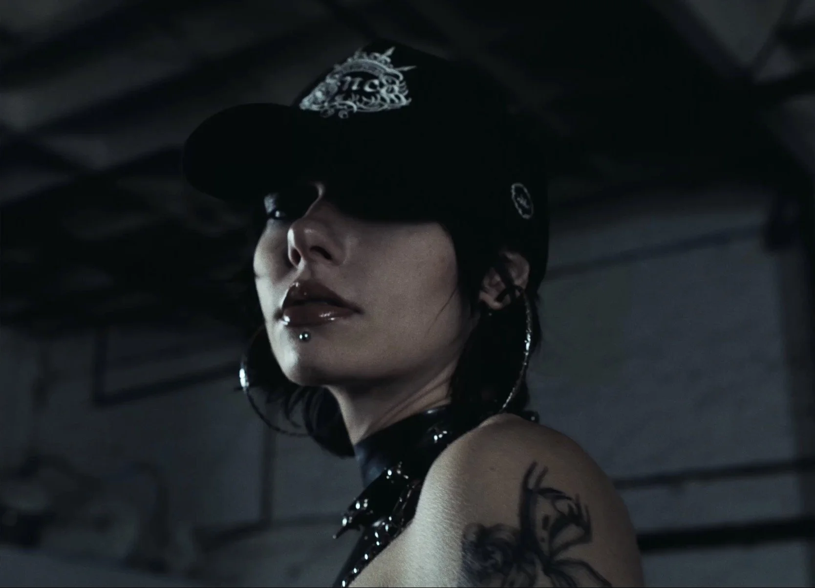 A woman with dark hair, tattoos, and piercings wears a baseball cap and large hoop earrings in a dark, industrial setting.