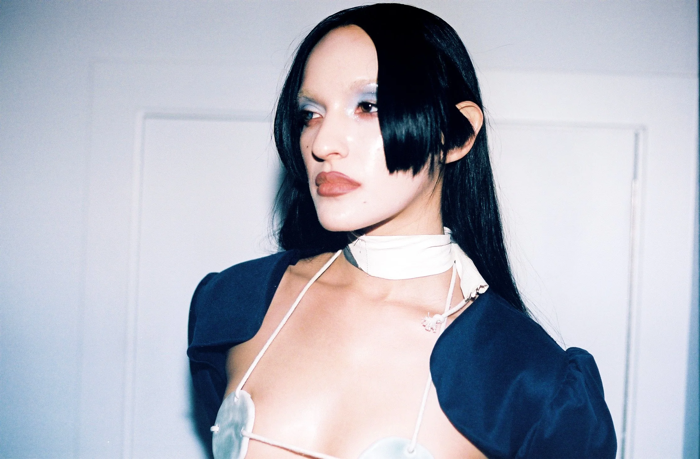 A woman with long black hair, blue eye makeup, and nude lipstick looking to the side. She is wearing a dark top with puffed sleeves, a white choker, and nipple covers connected by thin straps.
