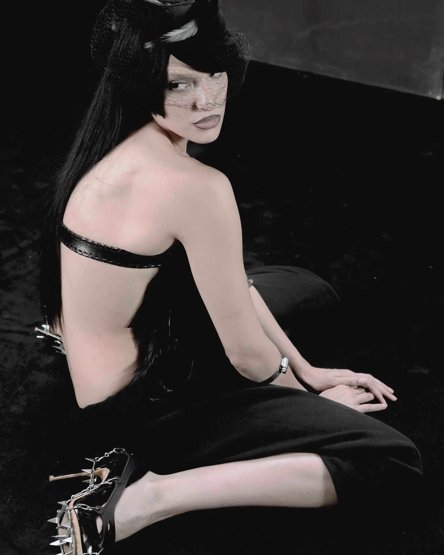 A woman with long black hair, wearing black spiked high heels, sits on the floor with her legs extended and turned to the side, looking over her shoulder at the camera. She is draped in black fabric, with a black strap across her back, and a black ne