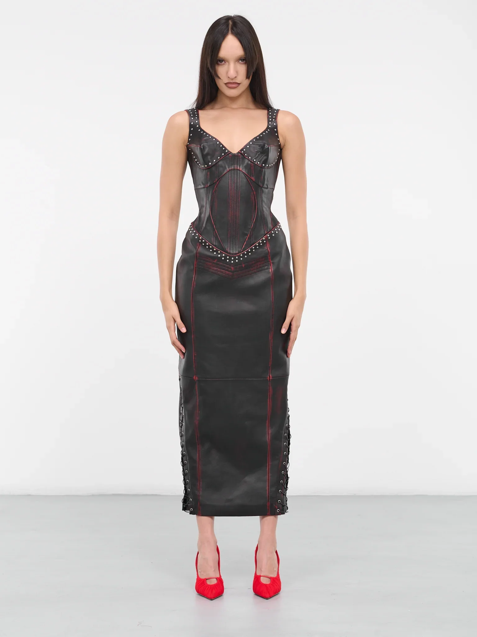 A woman standing against a plain white background wearing a black leather dress with red stitching, studded details, and lace-up accents on the sides, paired with red high heels.