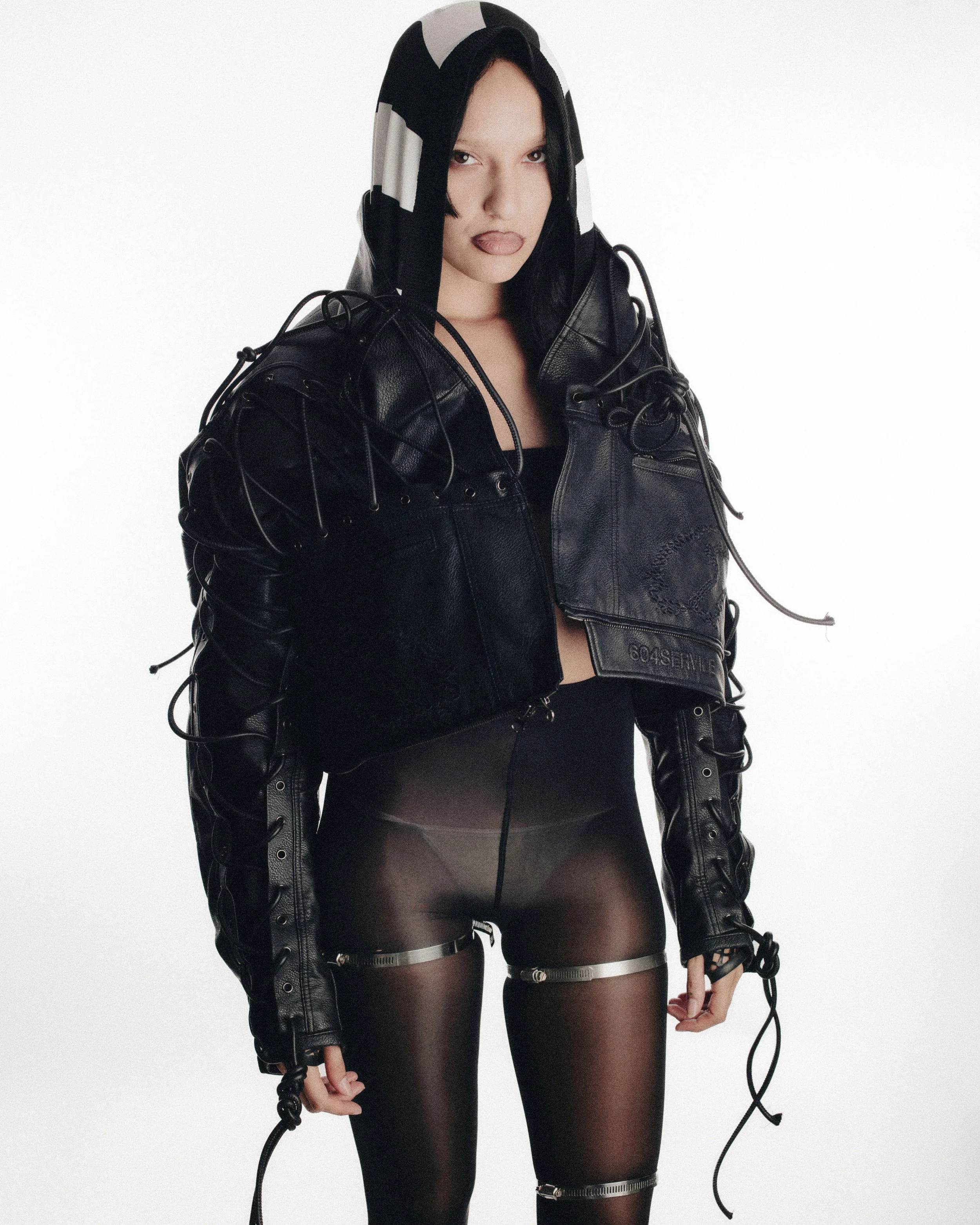 A woman wearing a black and white hood, a black leather jacket, and sheer black tights with zipper details. The jacket has laced embellishments, and she is making a playful facial expression with her tongue slightly out.