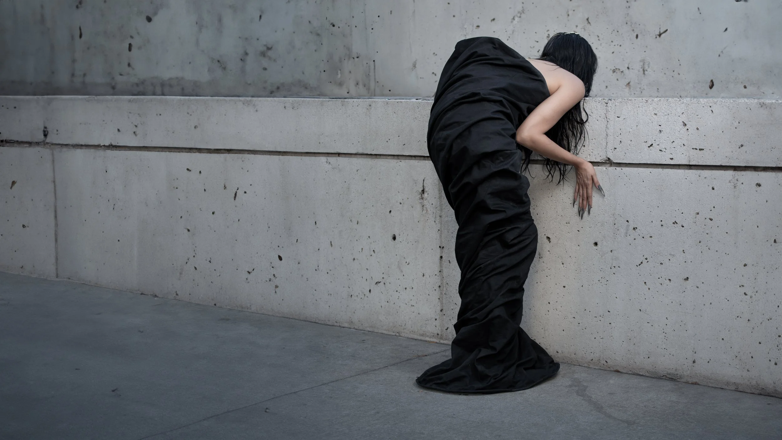 A person with long black hair, wearing a black sleeveless dress and black pants, is bent over with their head and hands against a concrete wall. The person's claws are visible and they are facing away from the camera.