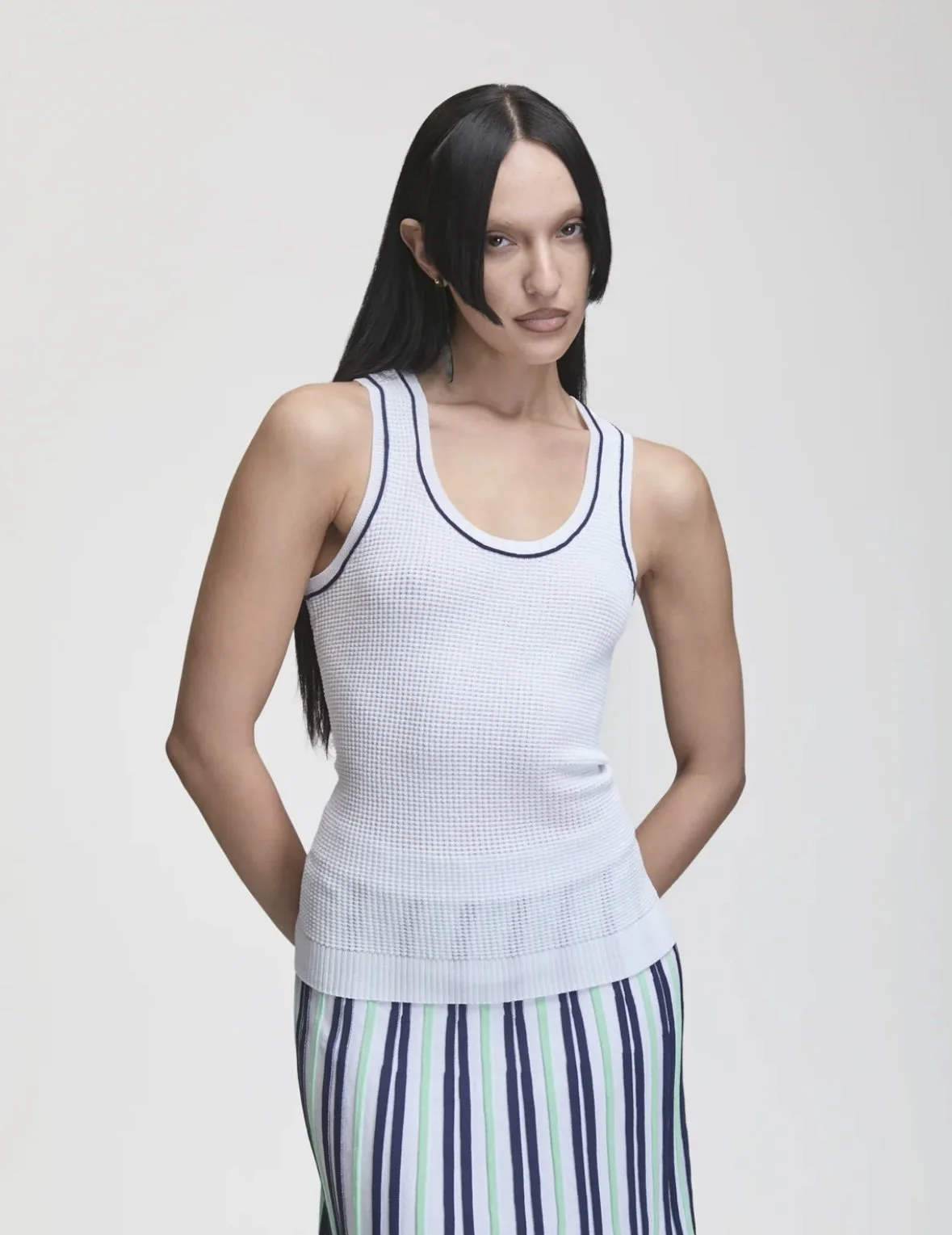 A woman with long black hair and wearing a sleeveless white top with blue trim, standing with her hands behind her back against a plain white background.