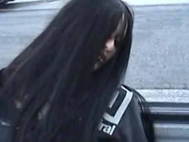 A woman with long black hair looking down, standing outdoors near a road
