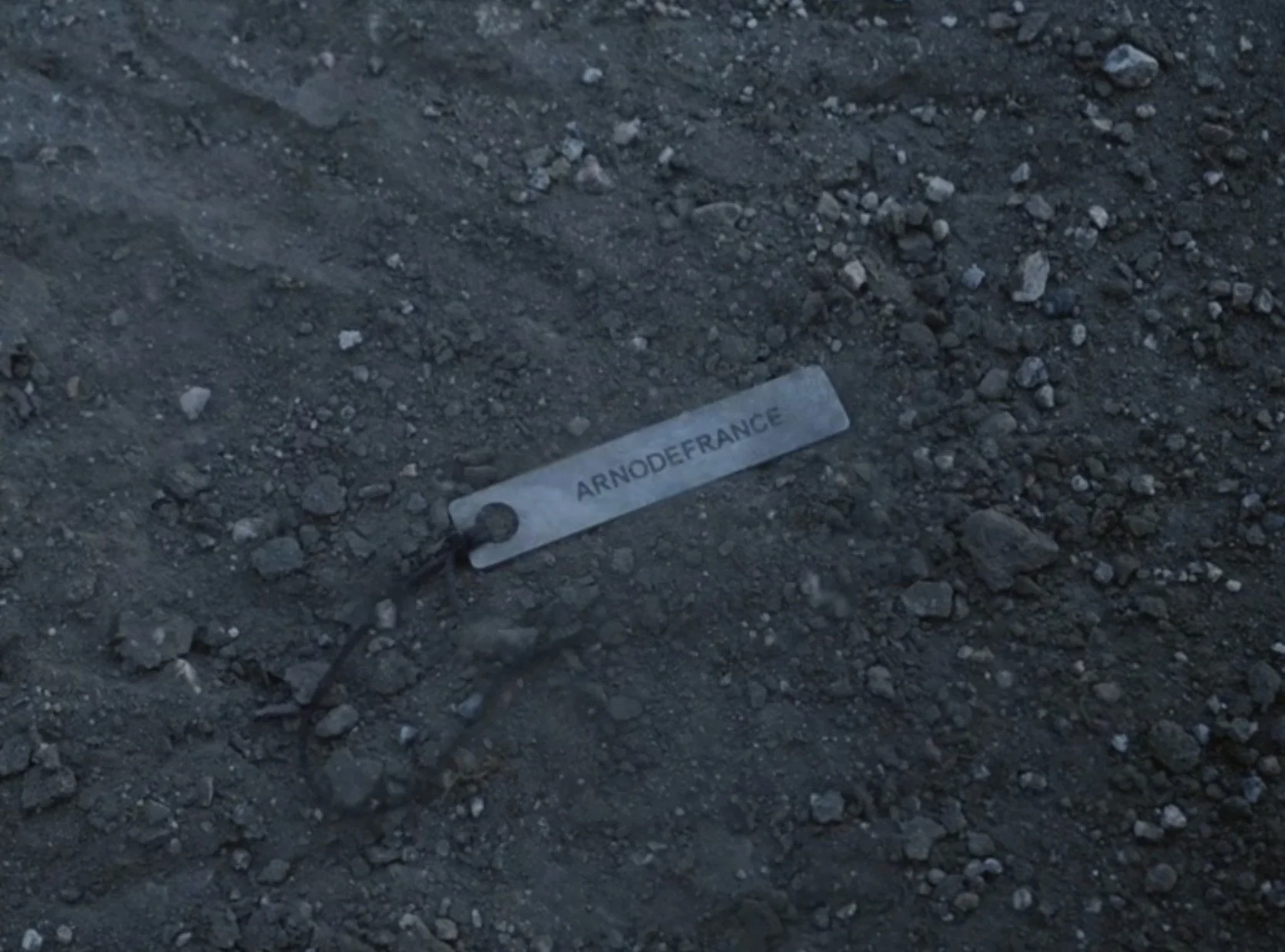 A metal tag with the word "ARNODEFRANCE" attached to a thin metal wire, lying on dark, rocky ground.
