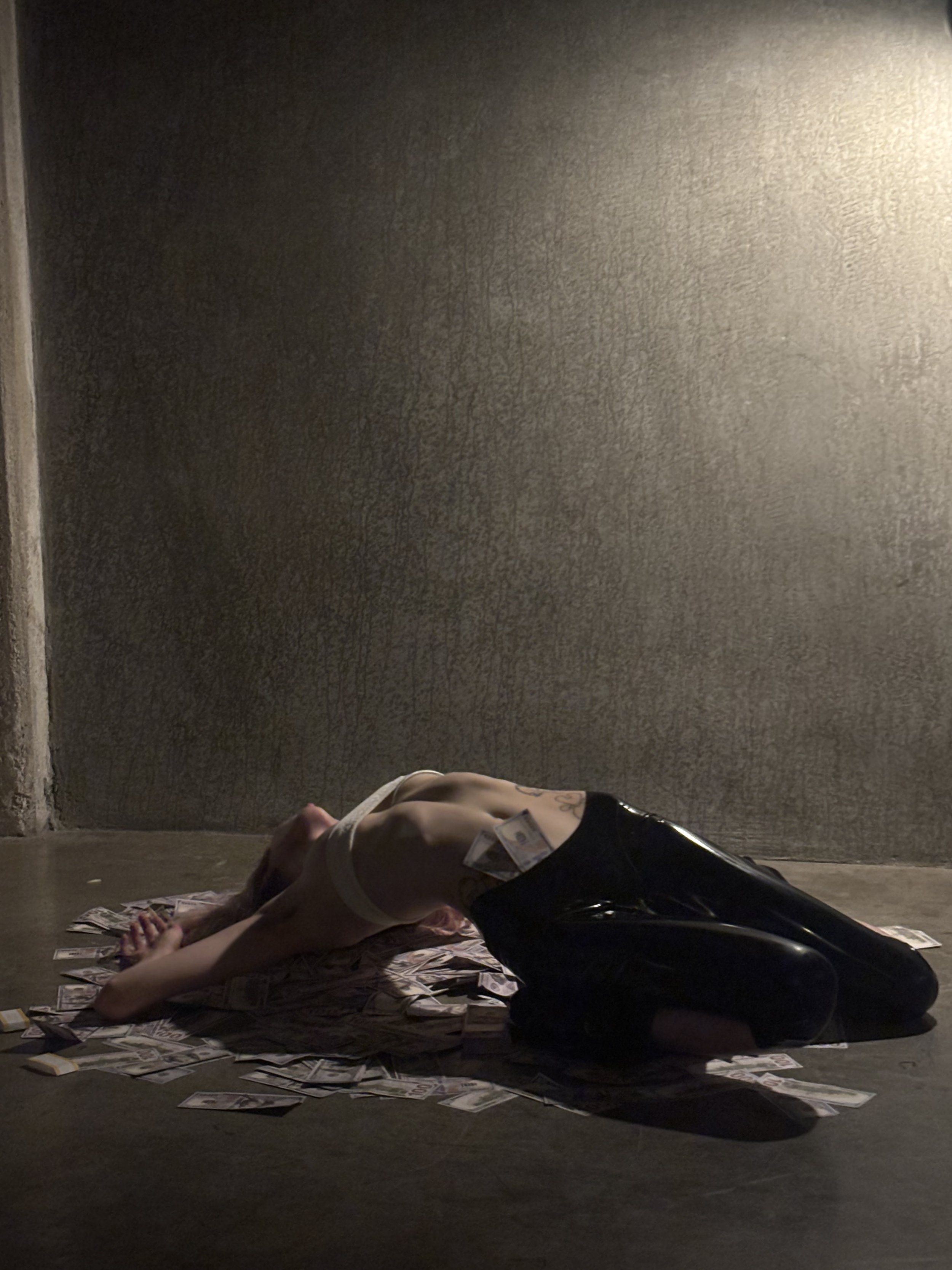 Person lying on the floor surrounded by scattered dollar bills in a dimly lit room.