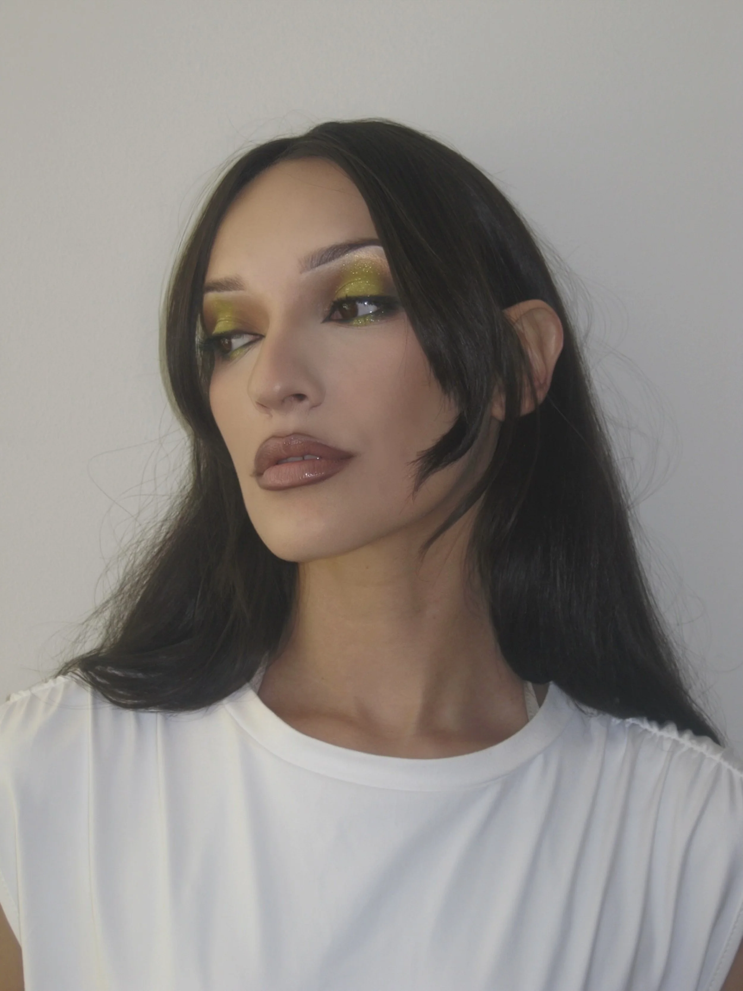 A woman with long black hair, wearing a white top and makeup with glittery green eyeshadow and nude lipstick, looking to the side against a plain background.