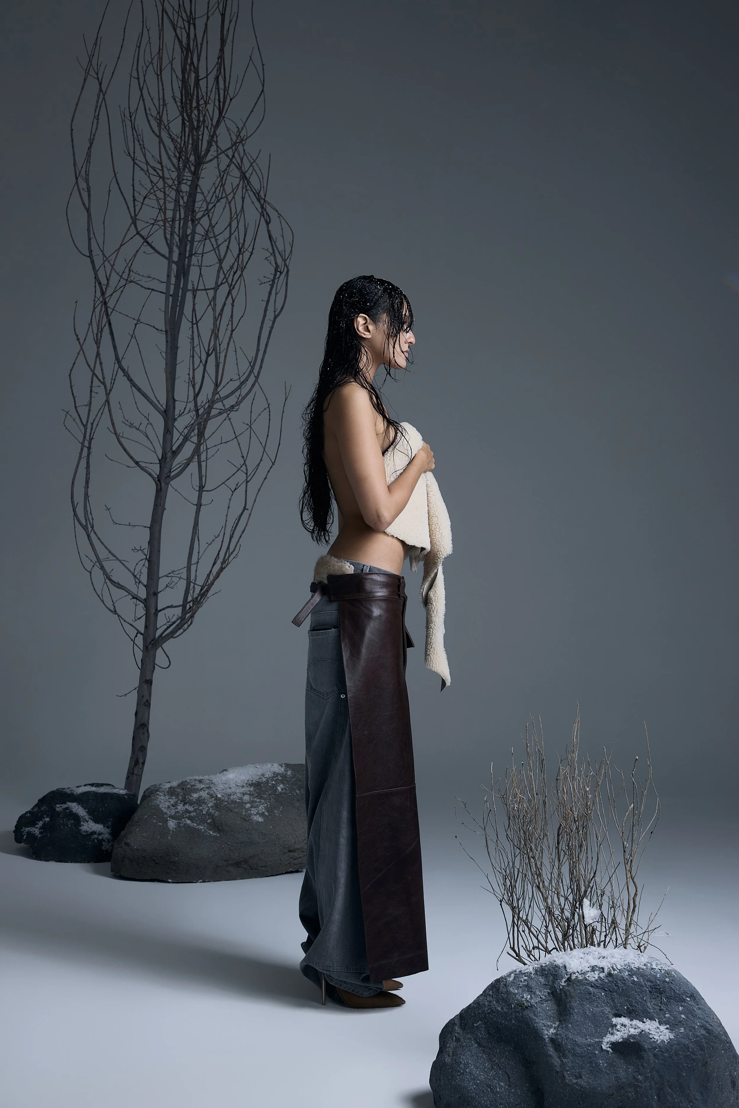 A woman with wet hair standing amidst a minimalist winter scene with rocks and dry trees, holding a towel against her chest.