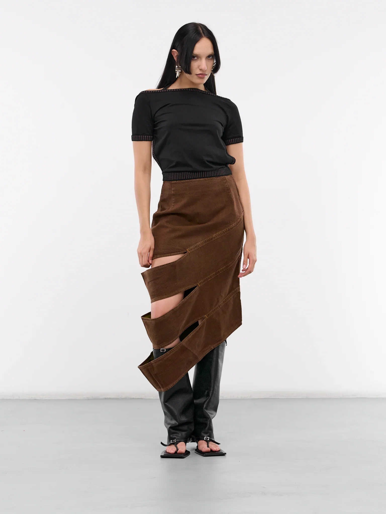 Fashion model wearing a black top and asymmetrical brown skirt with cutouts, paired with black open-toe sandals, standing against a plain white background.